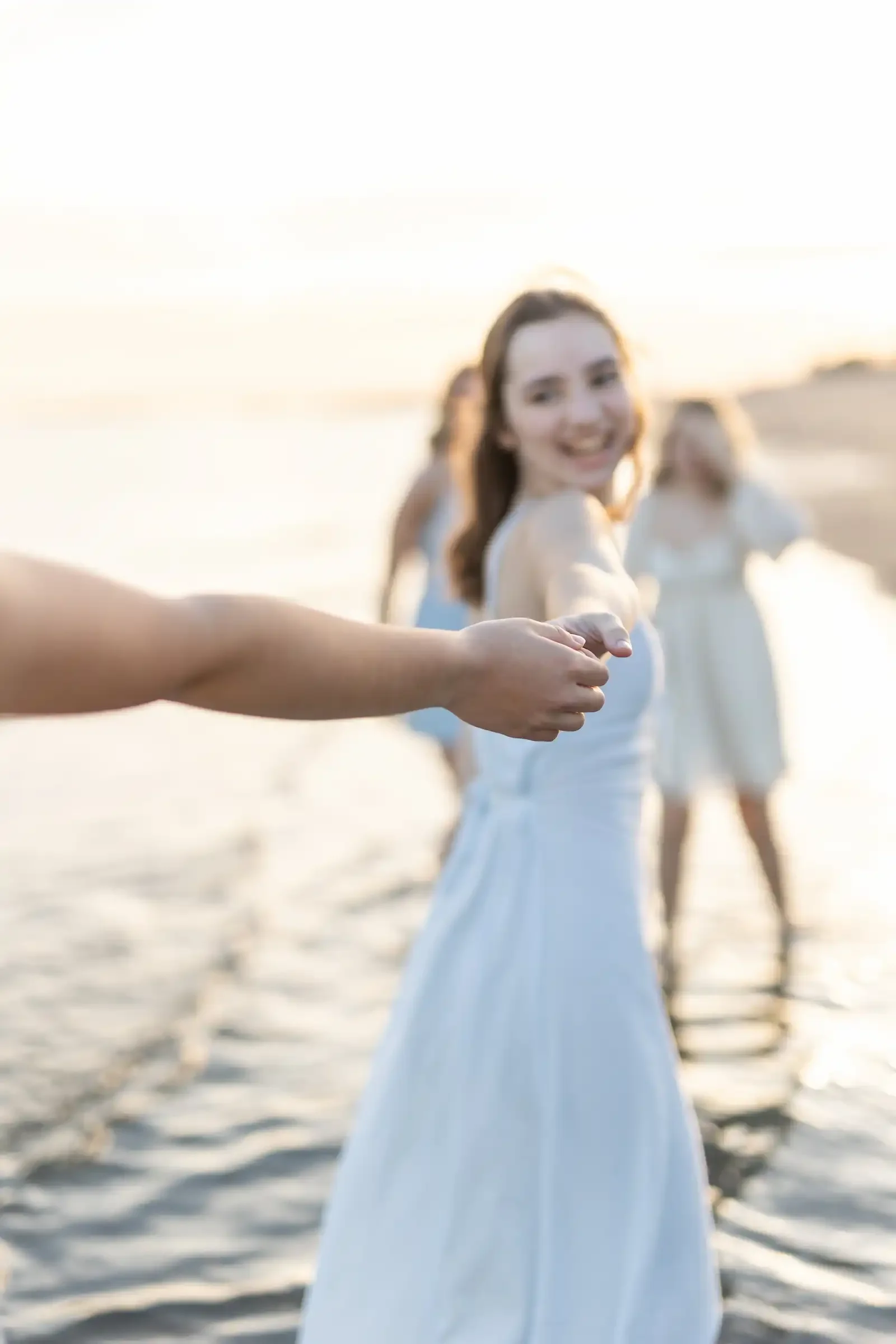 A group of women in white dresses holding hands on the beach during sunset.