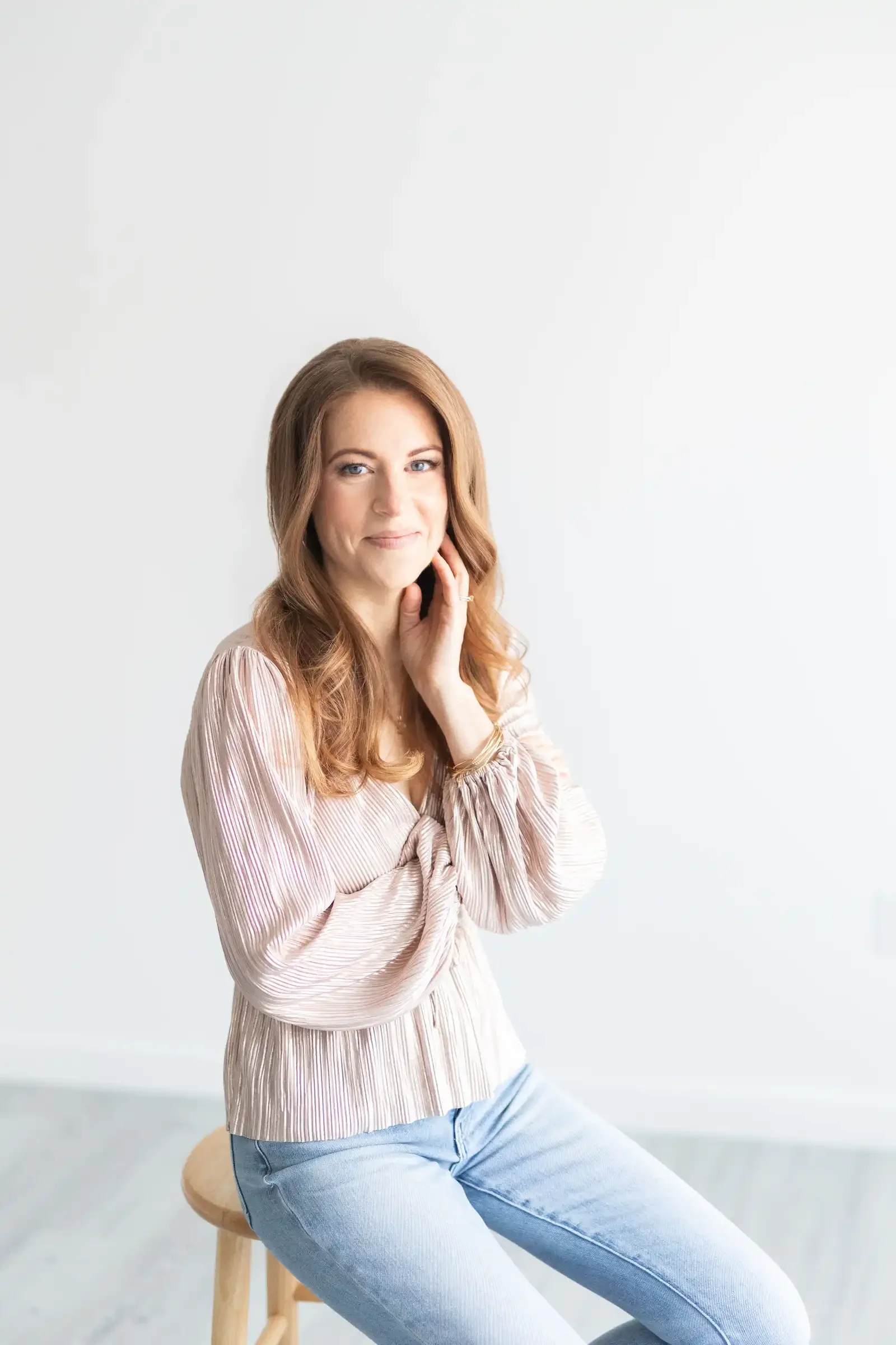 A woman with wavy, shoulder-length auburn hair sitting on a wooden stool against a plain white wall. She is wearing a light pink, pleated blouse and light blue jeans, with her left hand gently touching her face and smiling softly.