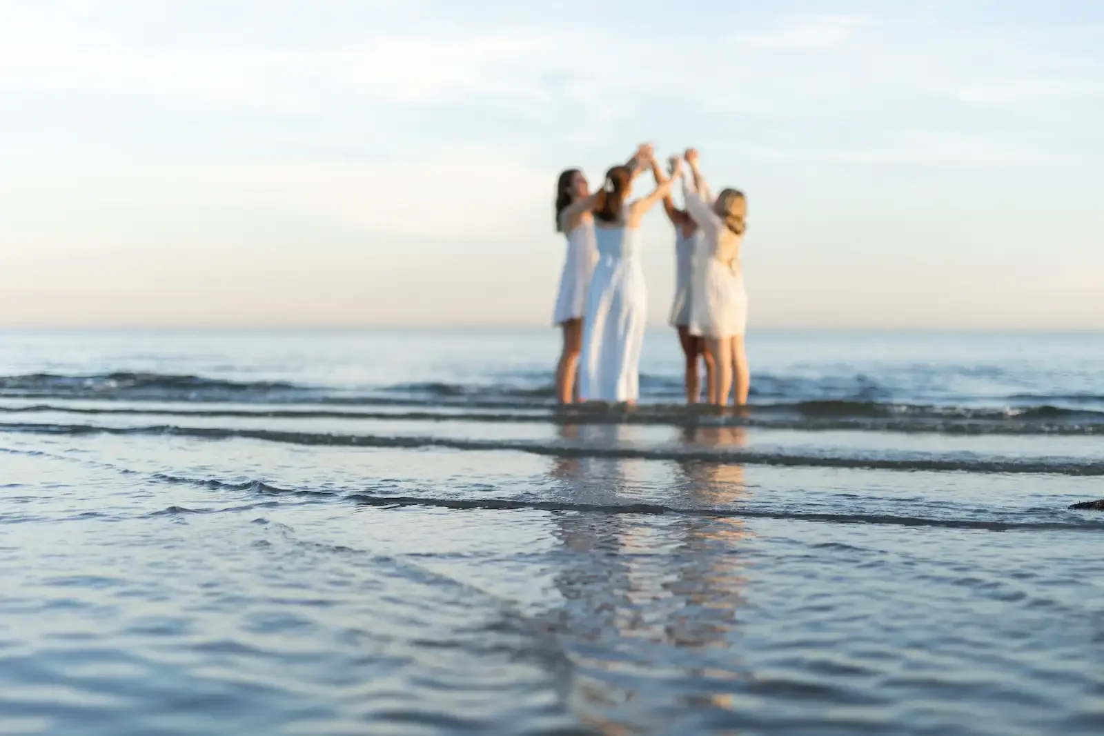 Four women in white dresses standing in shallow water at the beach, holding hands and forming a circle.
