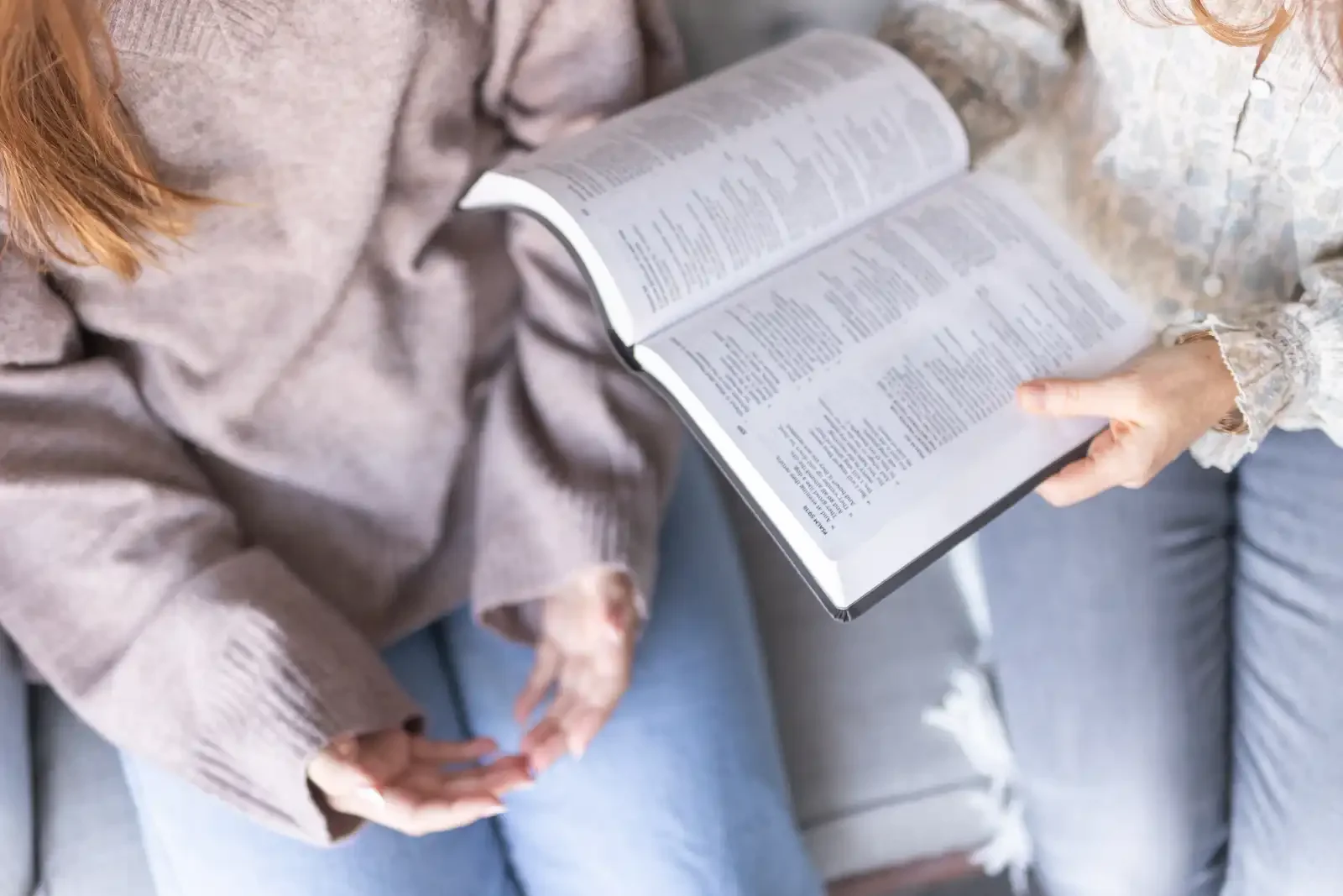 Person holding an open book while sitting on a light-colored sofa.