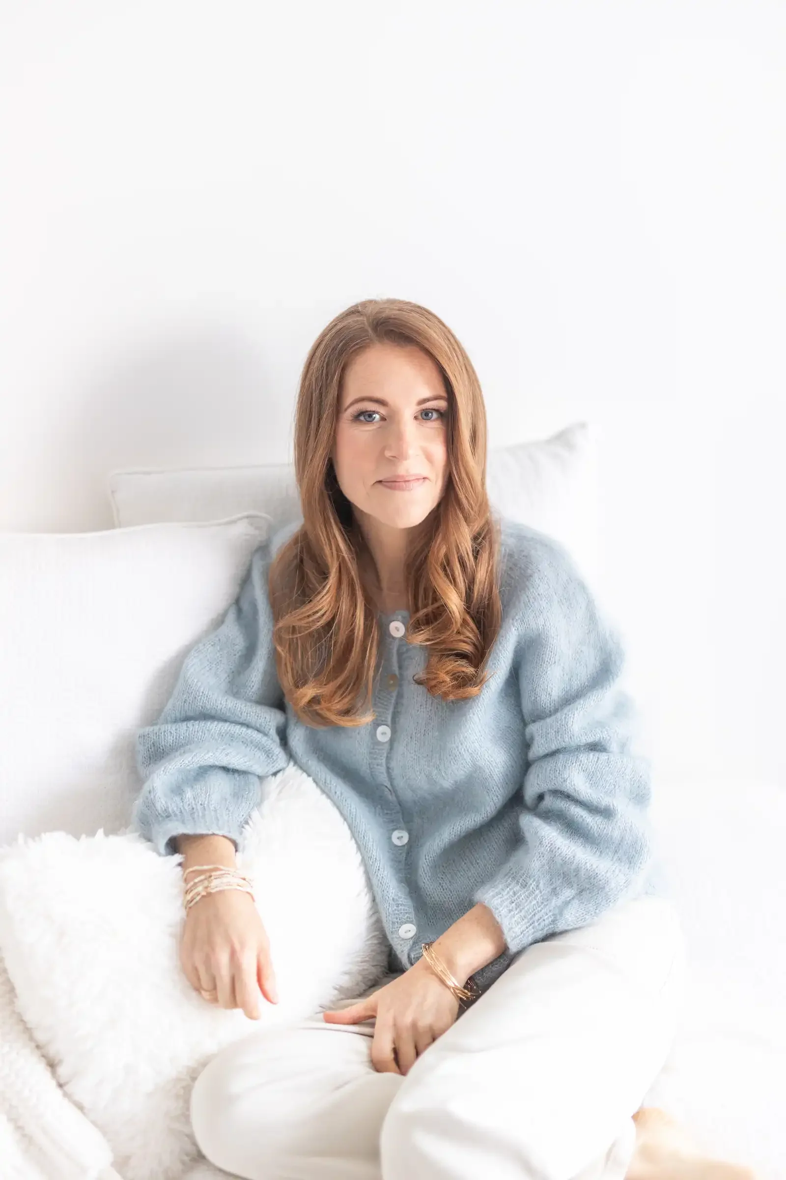 A woman with long reddish-brown hair sitting on a bed with white pillows and blanket, wearing a light blue cardigan and white pants, in a bright, minimal bedroom.