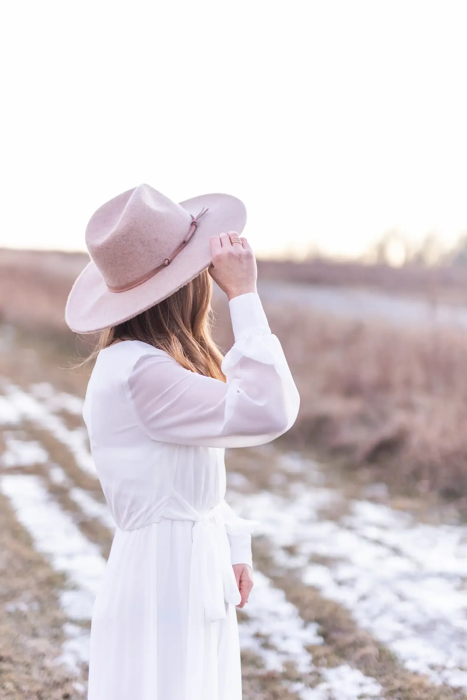 A woman in a white dress and a pink wide-brimmed hat stands outdoors on a winter landscape, looking into the distance with snow patches and a blurred background.