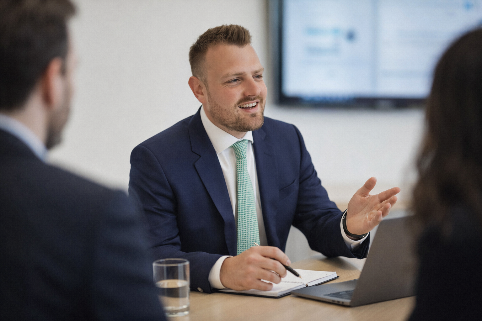 A man in a blue suit and green tie is speaking during a meeting, sitting at a table with a laptop, a notebook, a pen, and a glass of water in front of him. He appears to be explaining something to two colleagues, one with dark hair on the right and another with blurred features on the left.