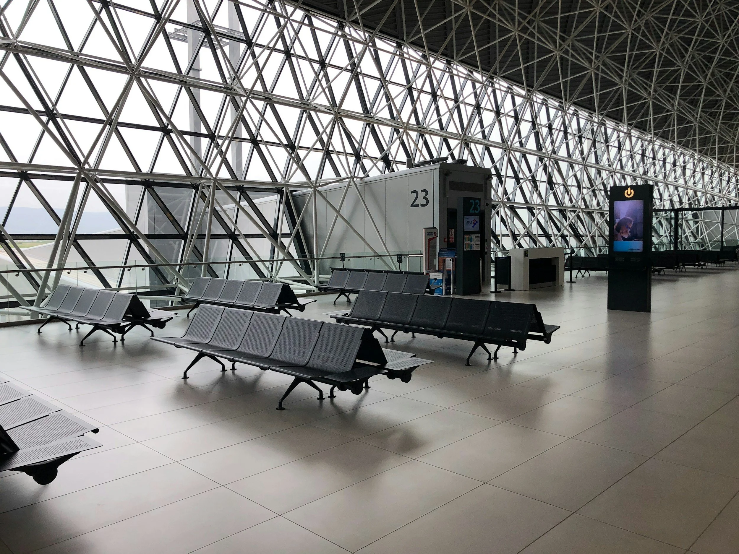 An empty airport waiting area with rows of gray seats, a large window with a grid-like metal framework, and a digital kiosk or screen near the gate 23.