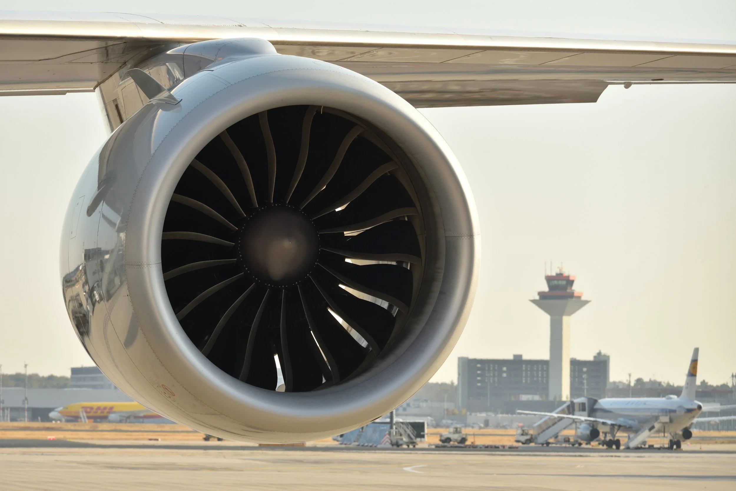 Close-up of a jet engine at an airport with an airplane and control tower in the background.