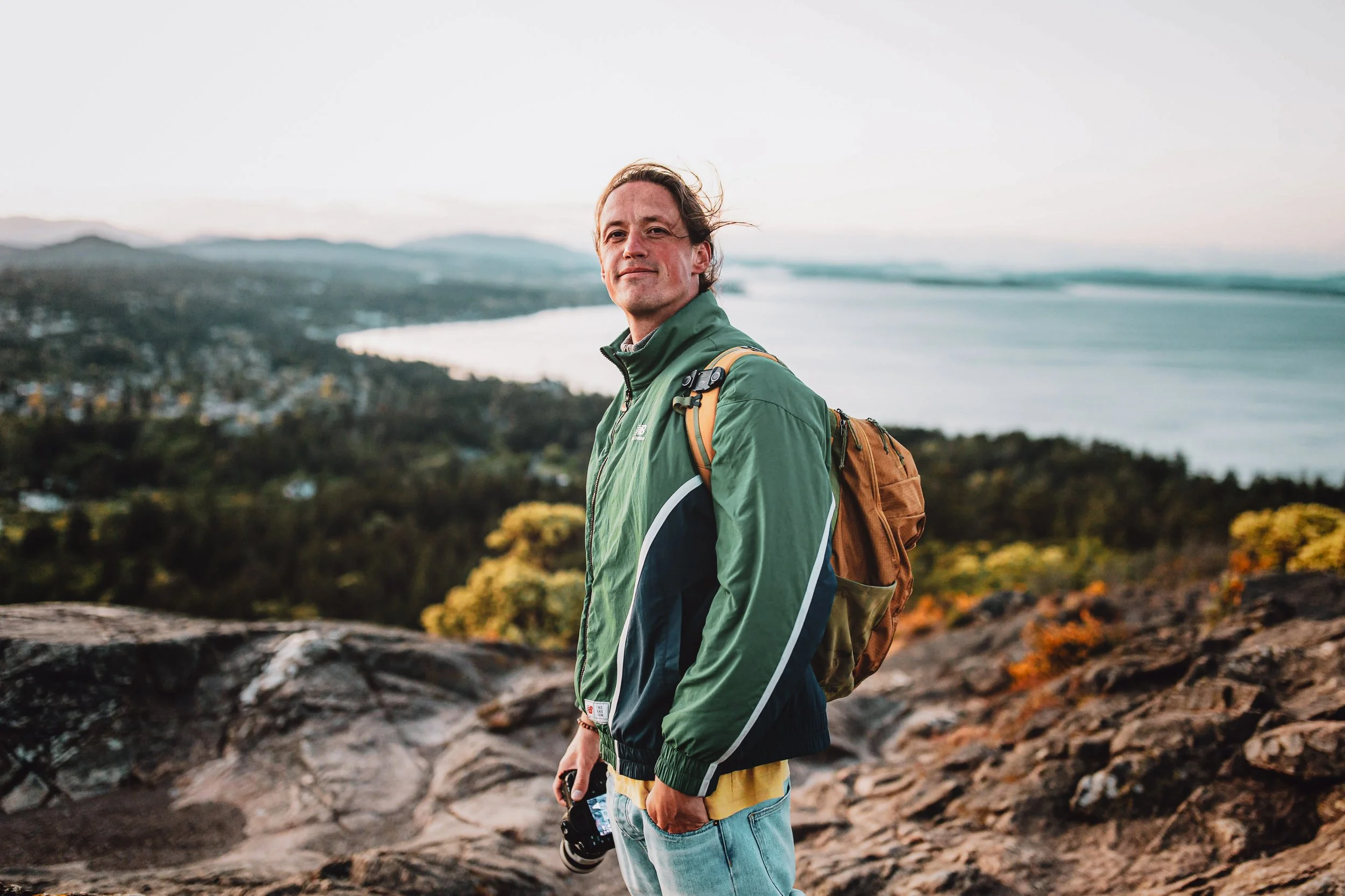 Man met een groene jas en een rugzak staat op een rotsachtige heuvel met uitzicht op een rivier en een bos in de verte tijdens zonsondergang.