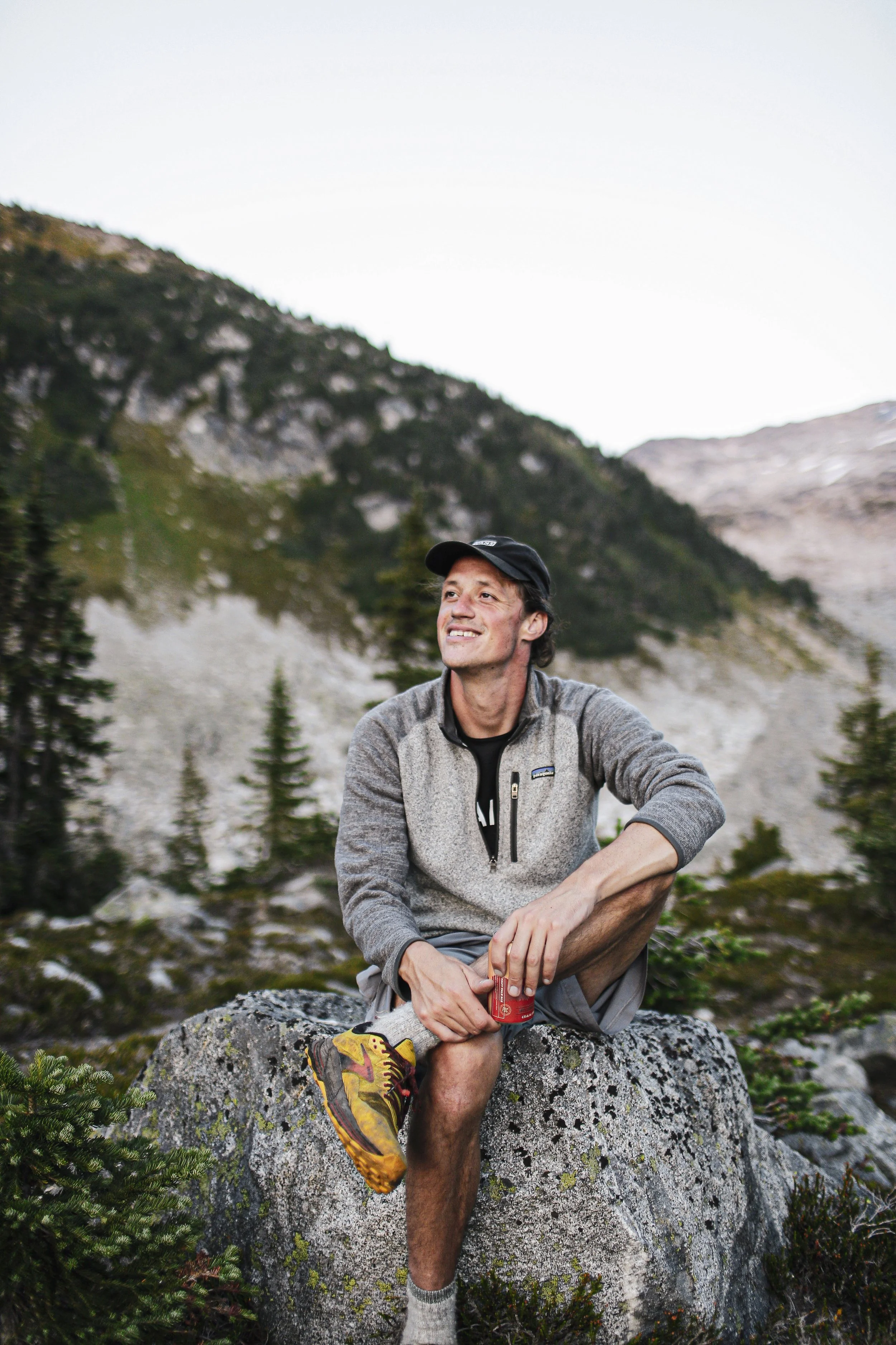 Man zittend op een rots in de natuur, omgeven door berglandschap en bomen, glimlachend en kermend met een drankje in de hand.