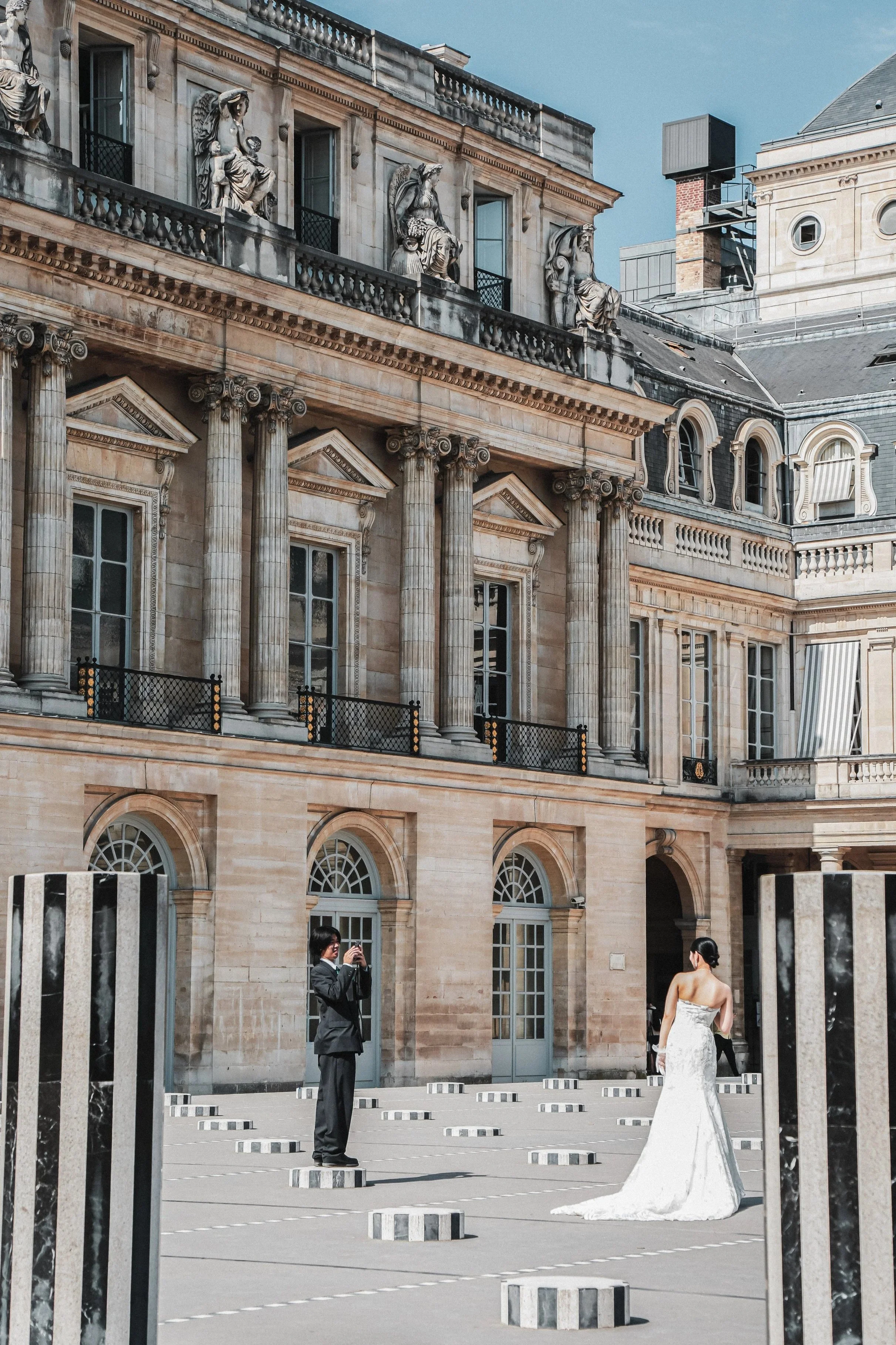 Een vrouw in een witte trouwjurk poseert voor een historicistisch gebouw met beelden en zuilen. Er is een man in een zwart pak die een foto van haar maakt. Ze staan op een openbare plek met ronde markeringen op de grond.