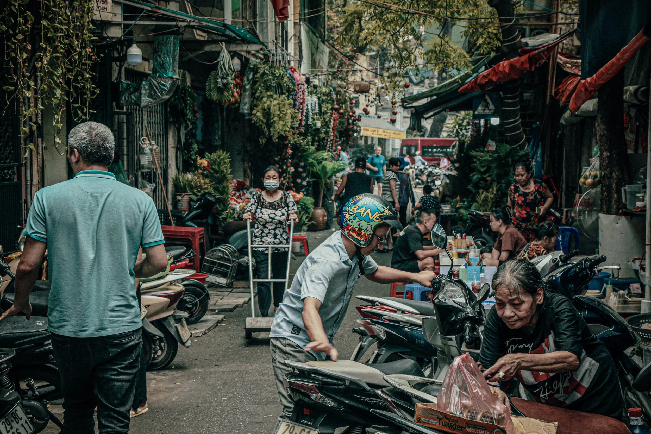 Een drukke straatmarkt met mensen die winkelen en praten, motorfietsen geparkeerd, bloemen en planten voor de winkels, en een rode bus op de achtergrond.