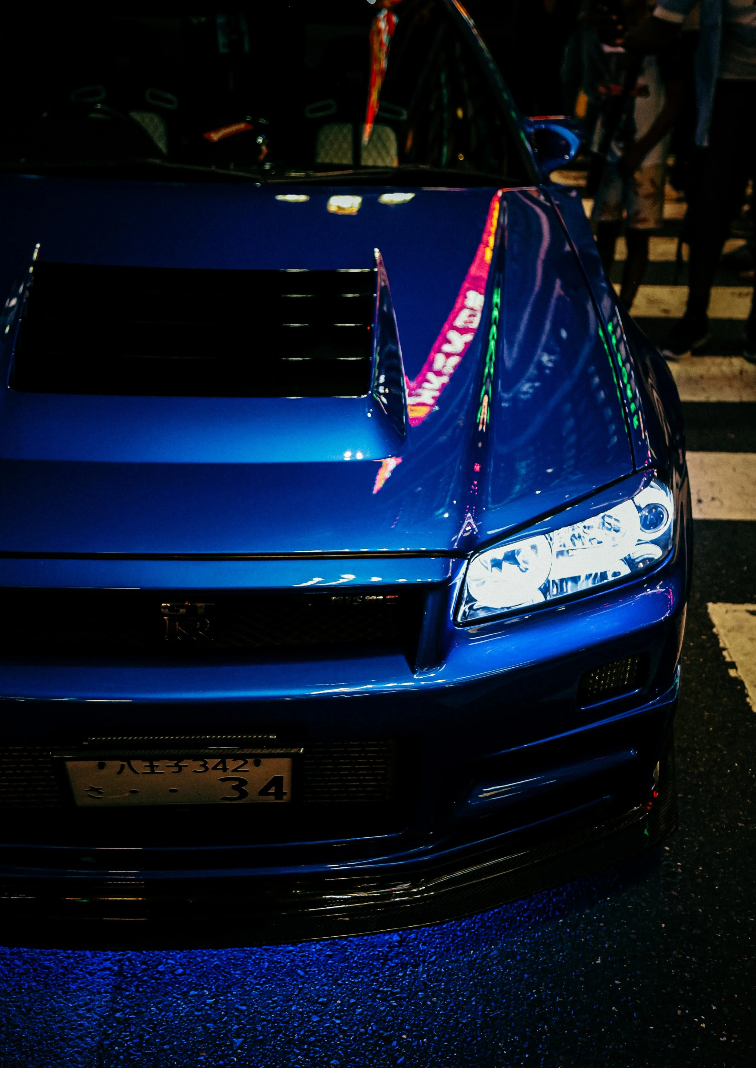 Een blauwe sportauto, een Nissan GT-R, geparkeerd op een stadsplein 's nachts, met neonverlichting reflecterend op het oppervlak.