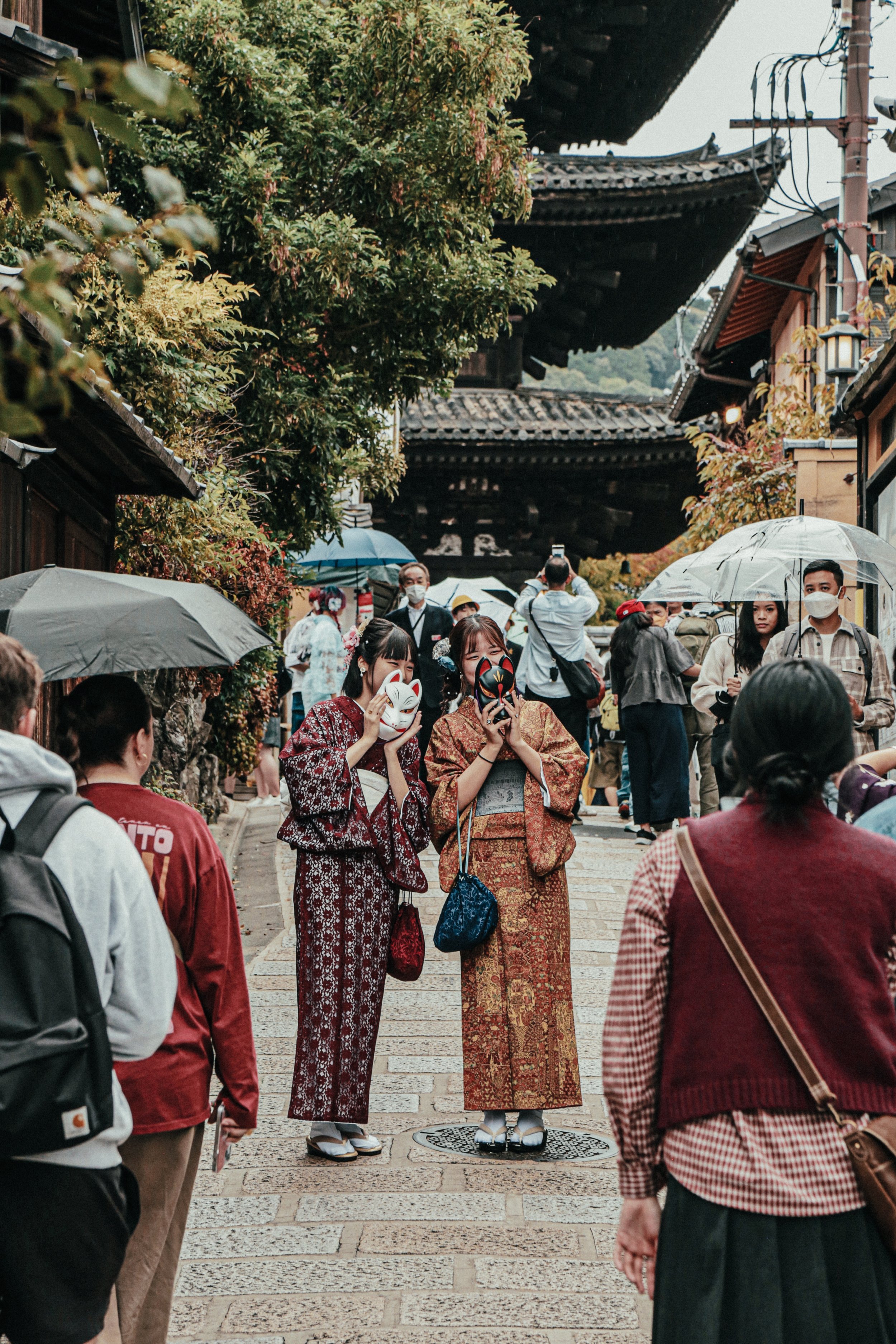 Twee vrouwen in traditionele Japanse kimono's poseert met maskers op in een drukke straat vol toeristen, met een oud Japans tempel gebouw op de achtergrond, en mensen met paraplu's.