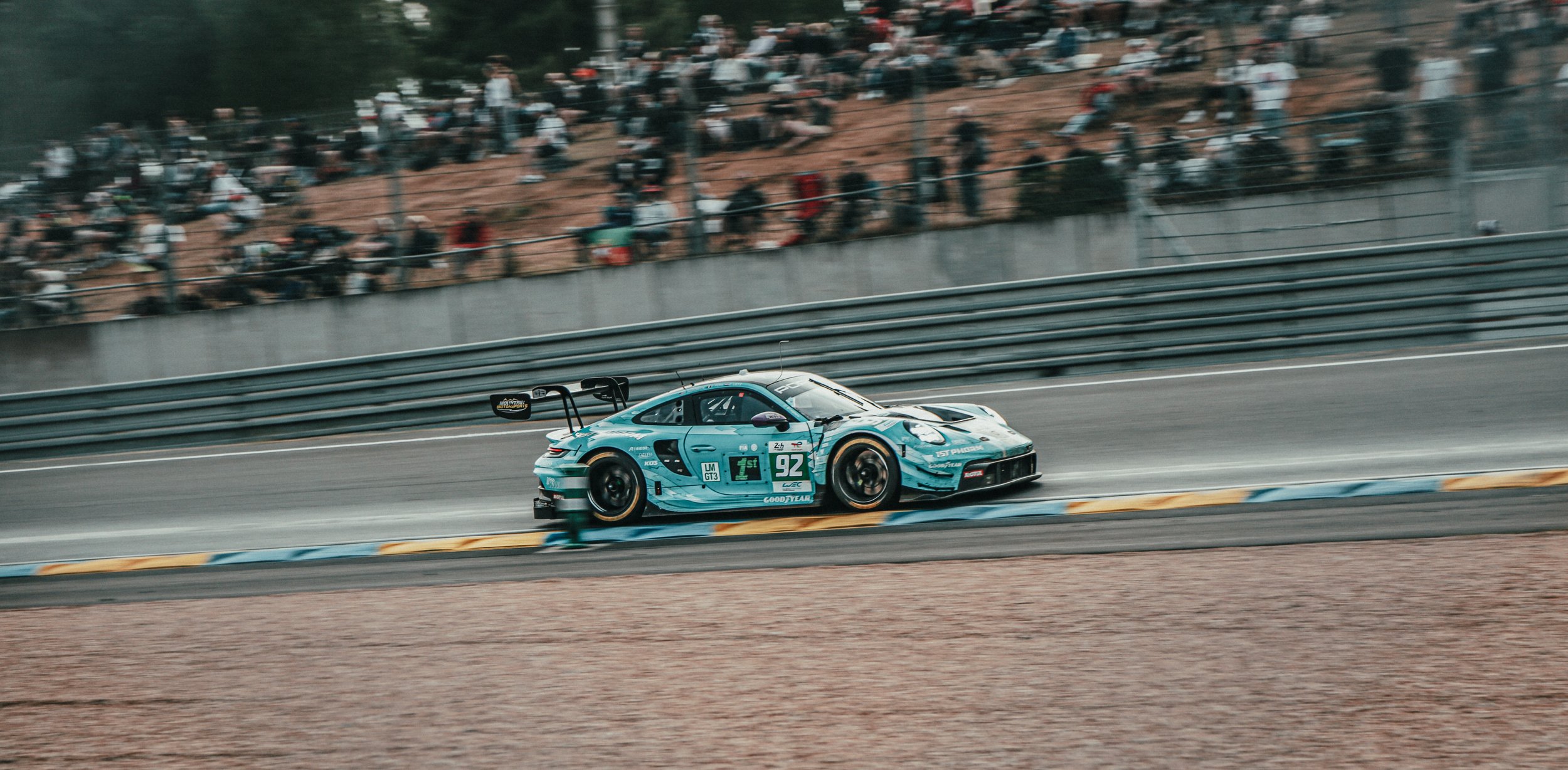 Een raceauto rijdt op een circuit met toeschouwers op de heuvels op de achtergrond.