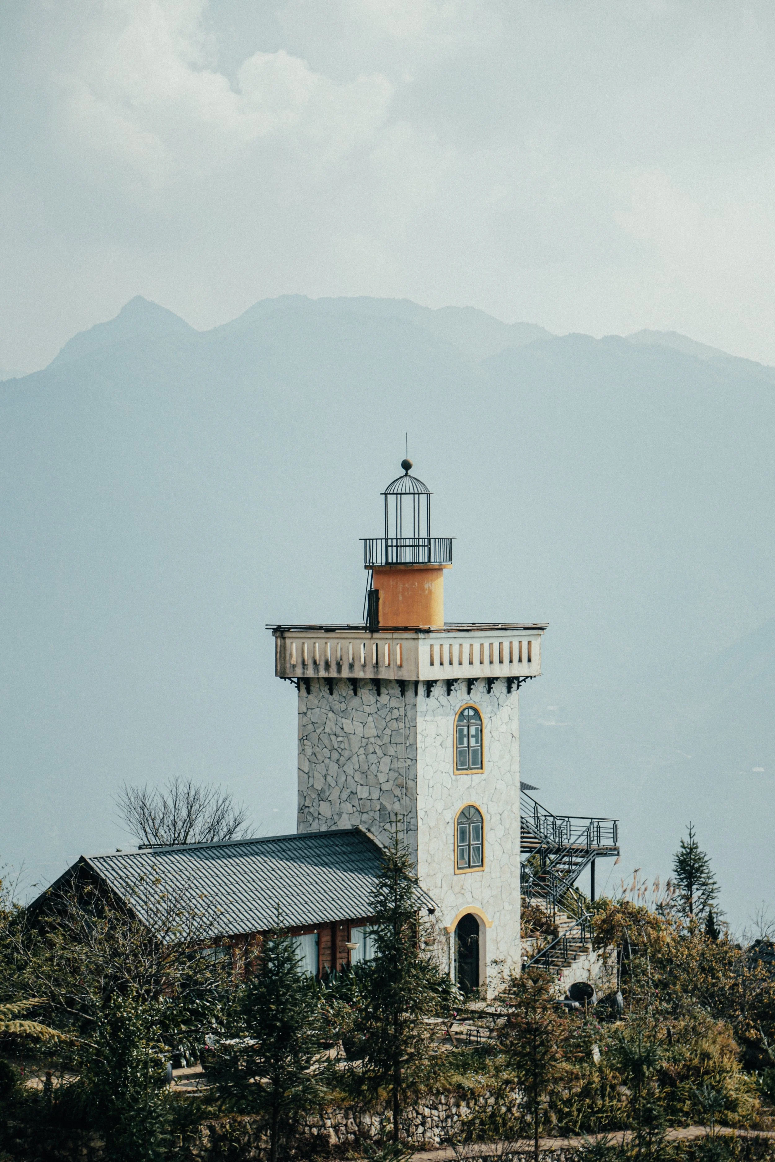 Een toren met een vuurtorenlamp en een balkon, omgeven door bomen en met bergen op de achtergrond.