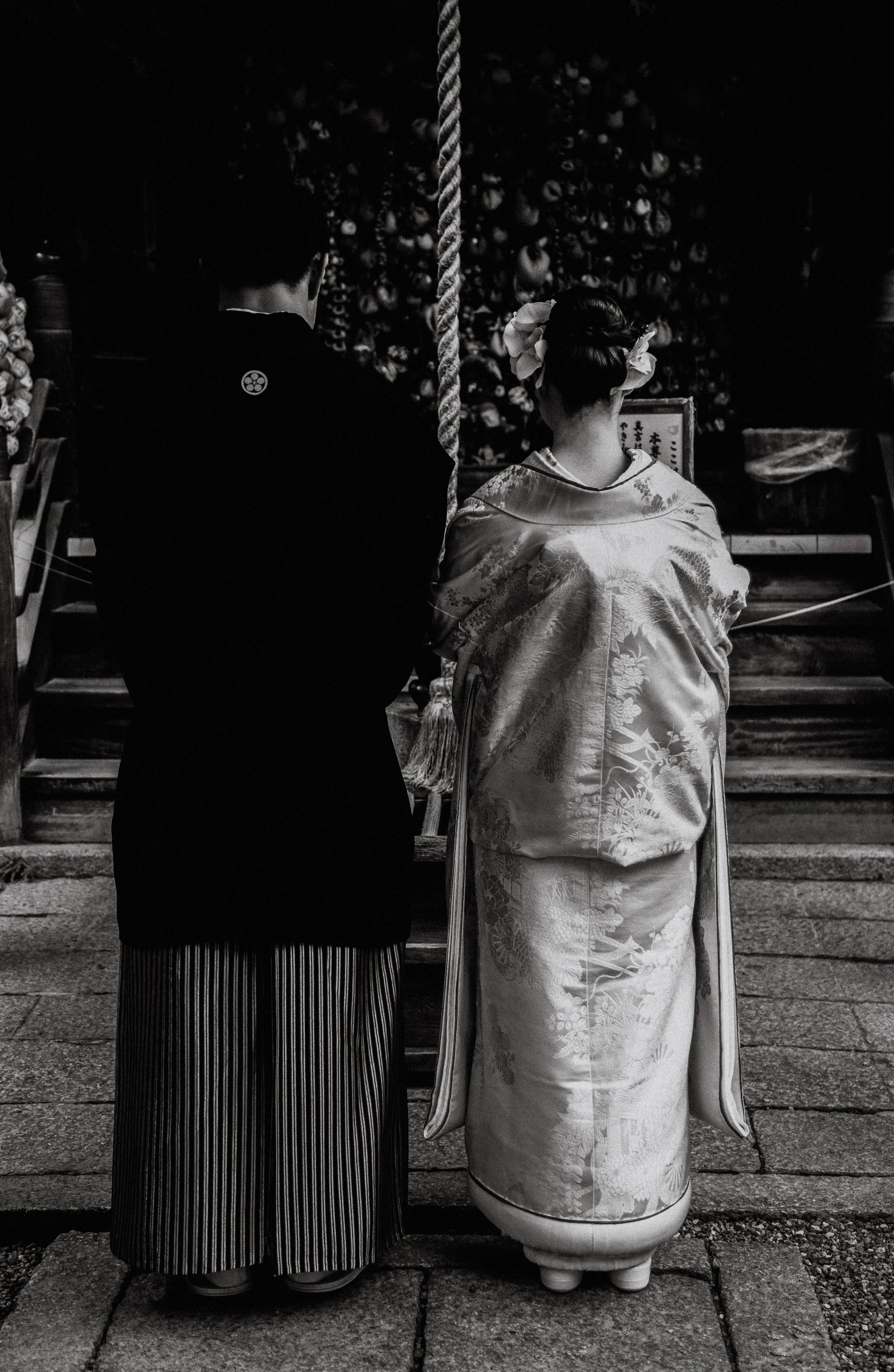 Twee mensen gebogen voor een altaar in een traditionele Japanse ceremonie, een man en een vrouw in kimono, gezien van achter. Kostuums en achtergrond met religieuze ornamenten.