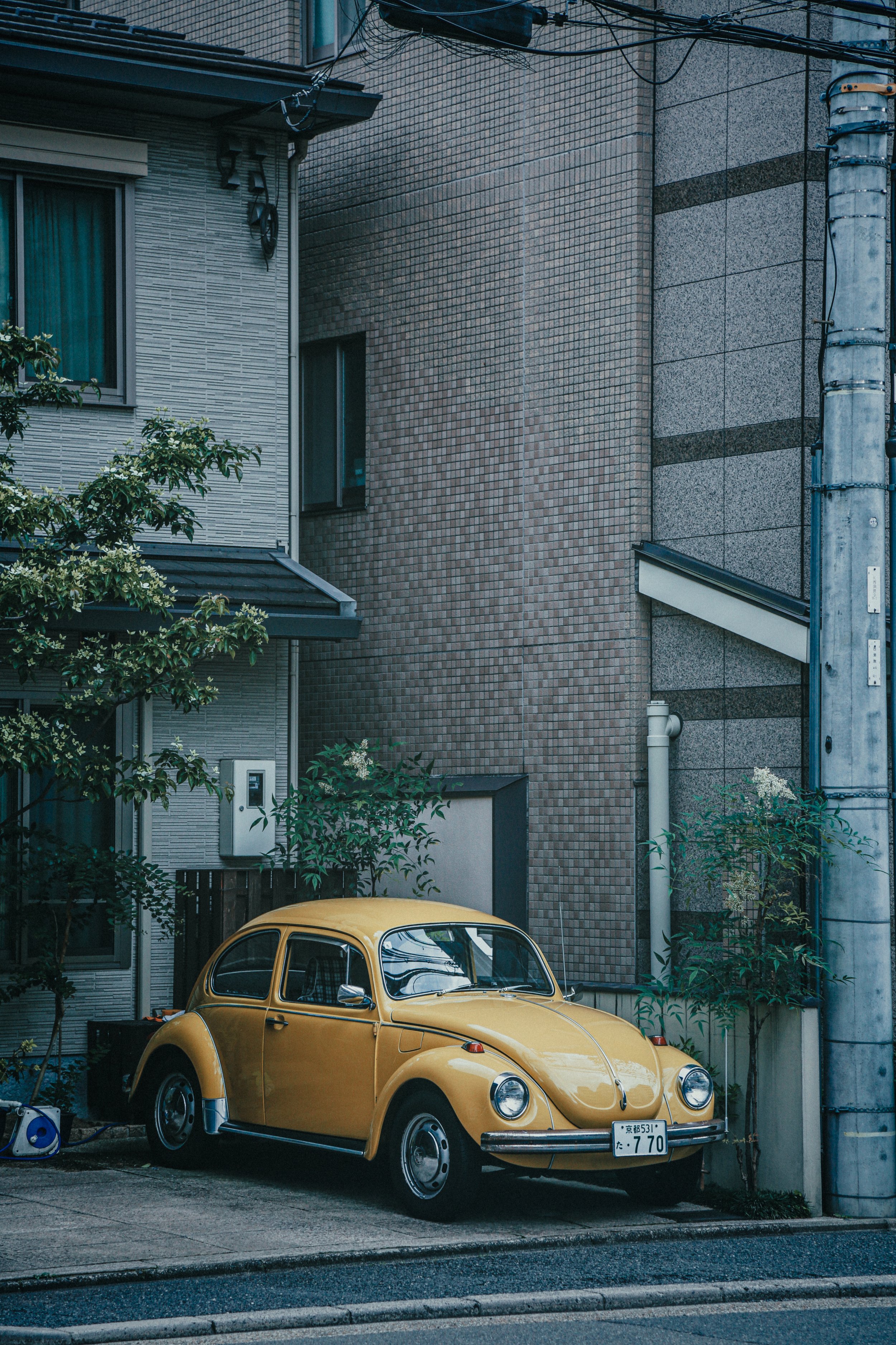 Een oude gele Volkswagen Kever geparkeerd naast een gebouw met bakstenen en betonnen muren, omgeven door groene bomen.