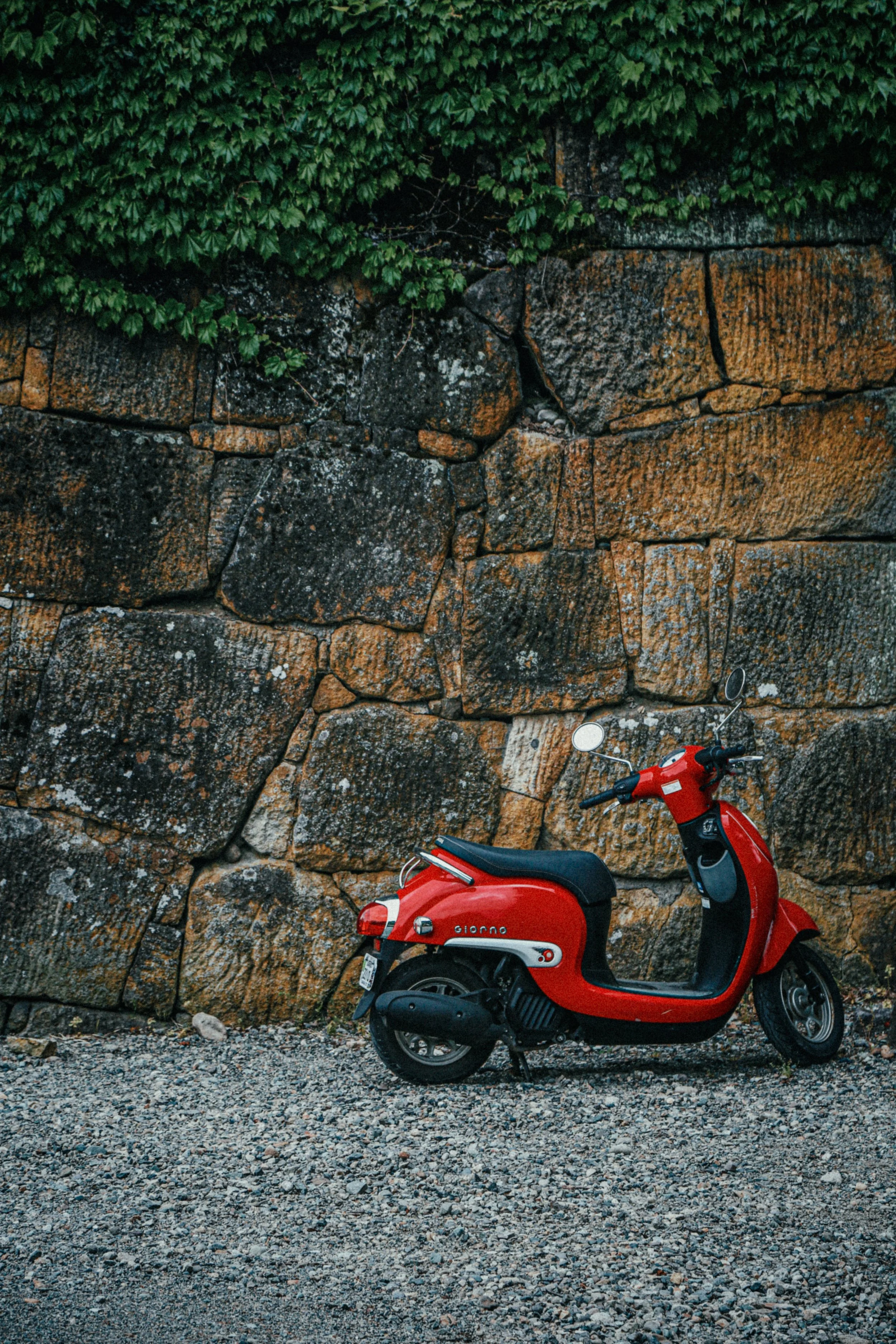 Een rode scooter geparkeerd voor een stenen muur met groene begroeiing bovenaan.