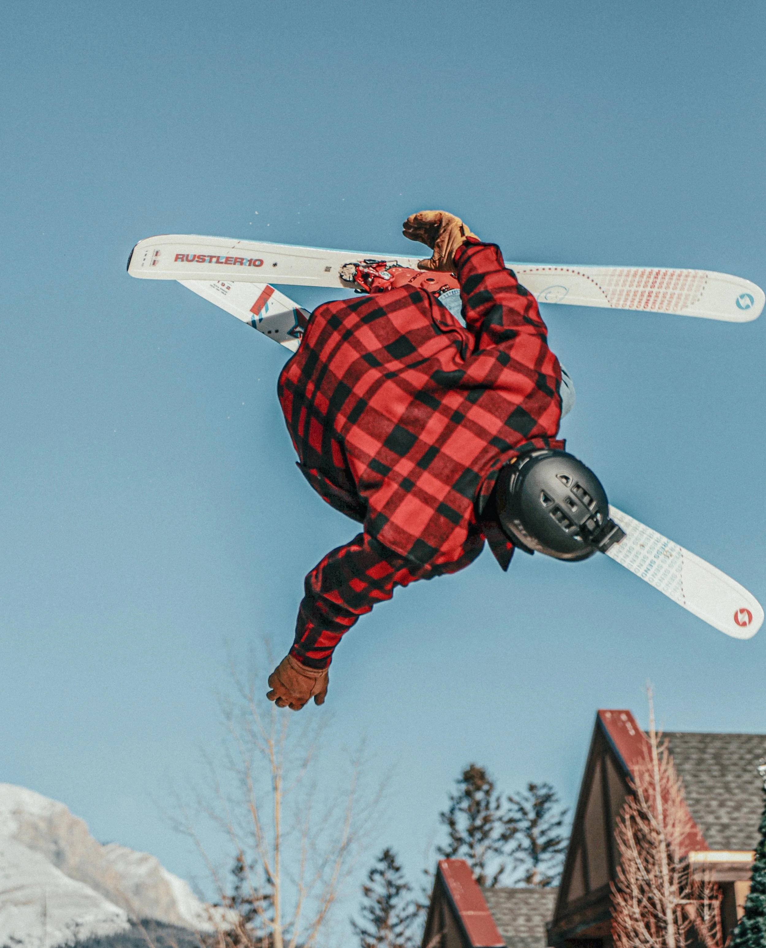 Skiër voert sprong in de lucht uit, met blauwe hemel en daken van huizen op de achtergrond.