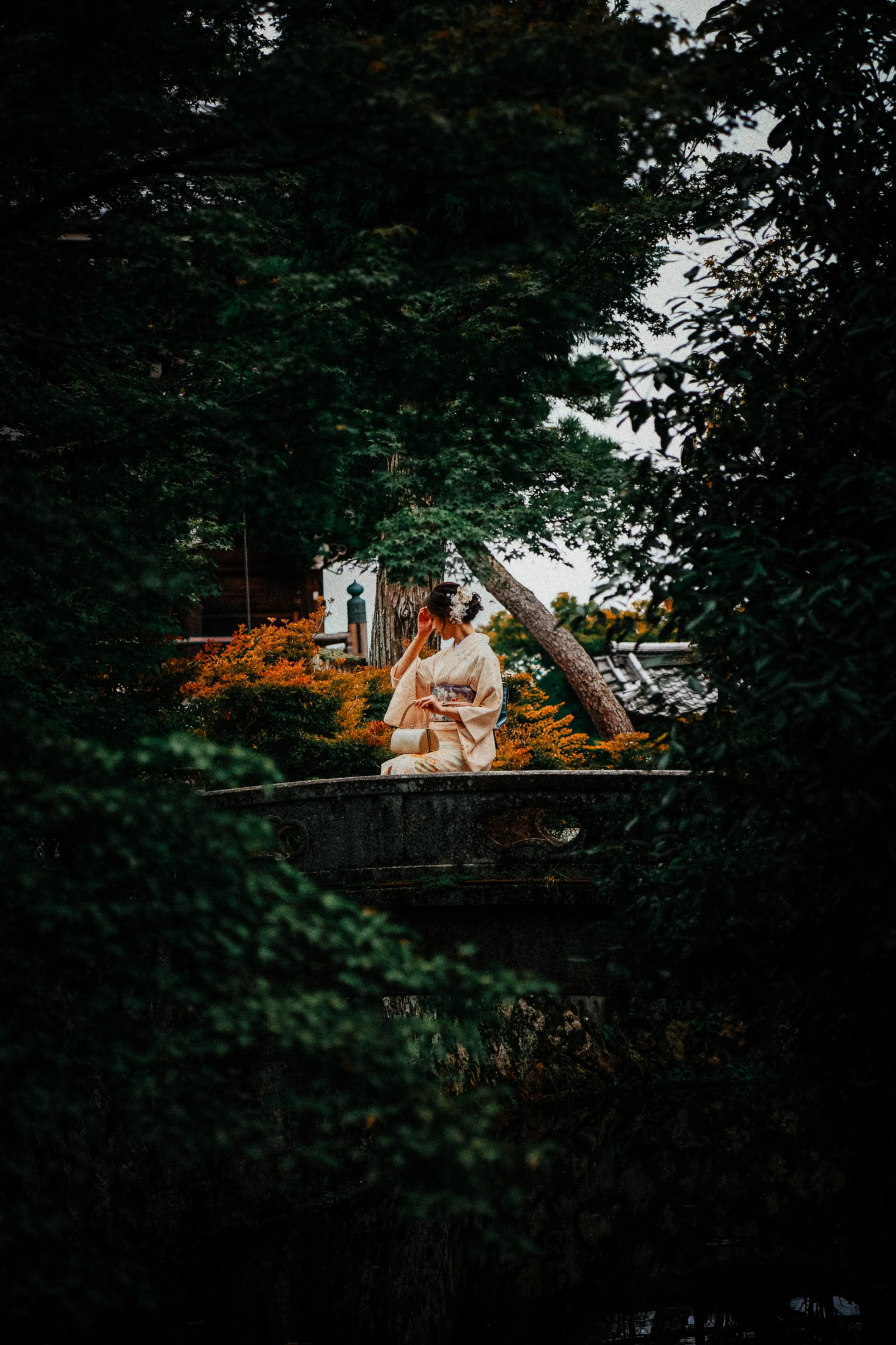 Vrouw in kimono zit op een brug in een Japanse tuin, omgeven door groen en oranje struiken.