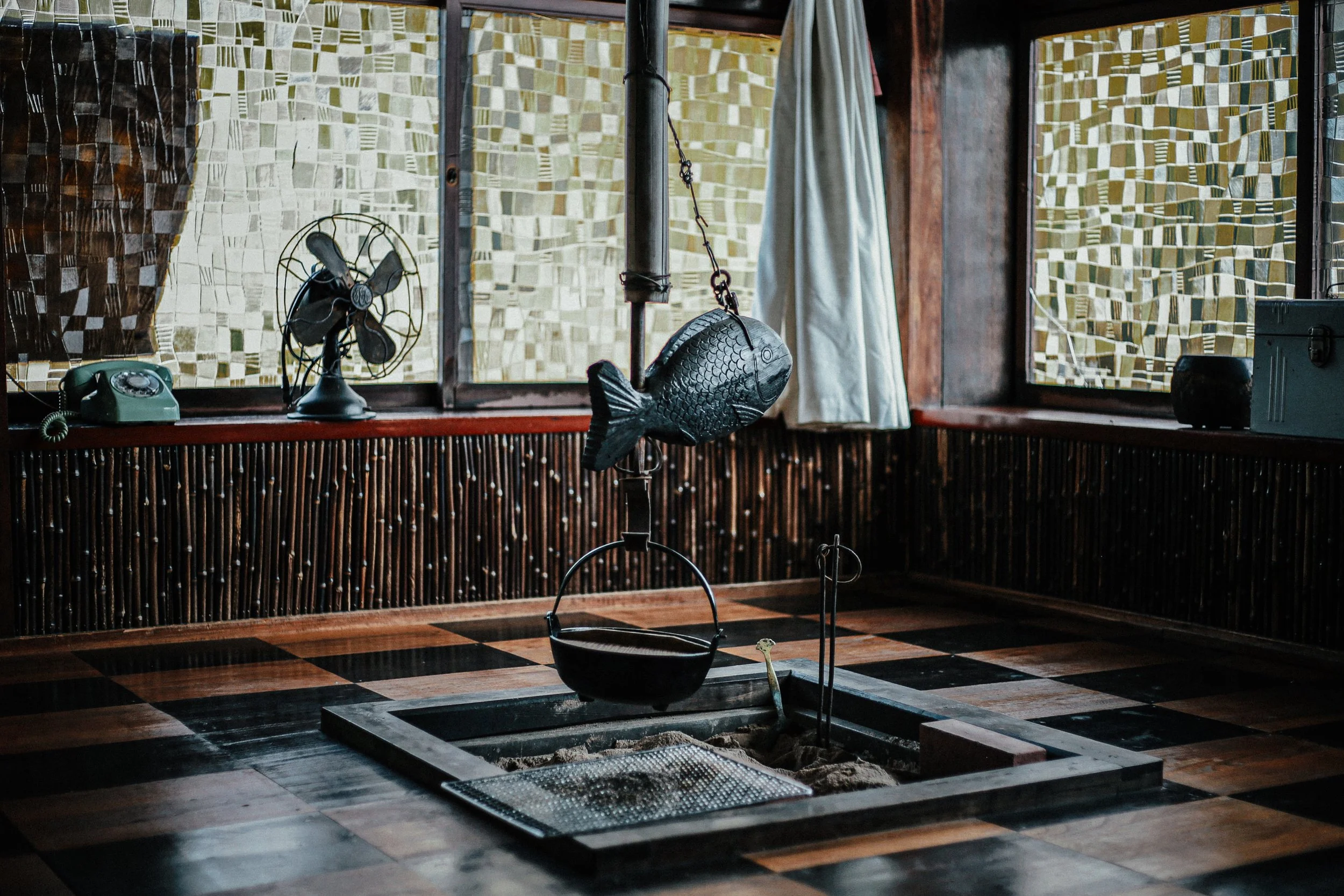 Interieur van een kamer met vintage decor, inclusief een oude telefoon, een kleine ventilator, en hangende decoratie in de vorm van een vis. De kamer heeft grote ramen met cassette-deuren en houten omlijstingen, en een vloer met een zwart-wit tegelpa
