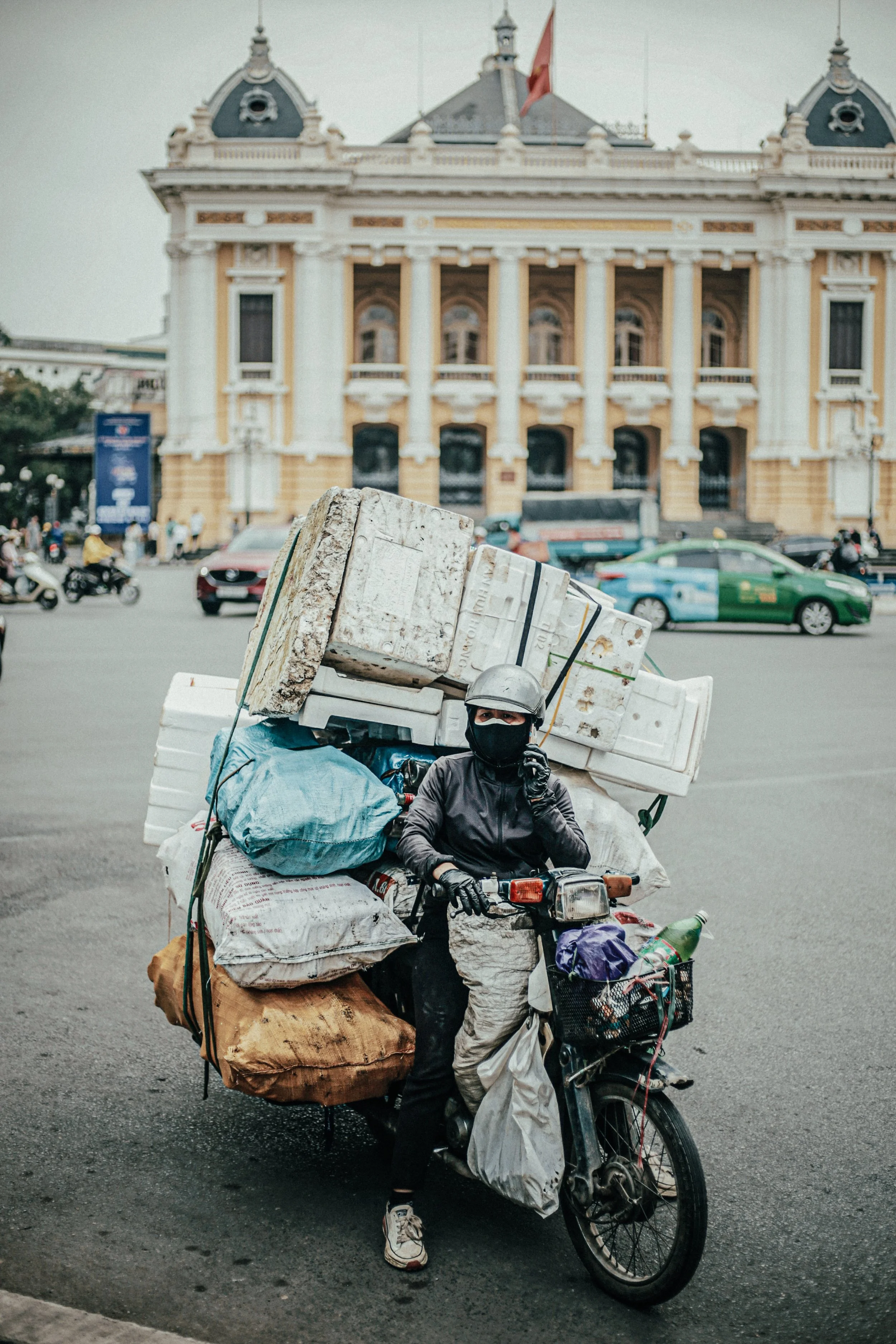 Man op een motorfiets volgeladen met spullen op straat, met een historisch gebouw op de achtergrond.