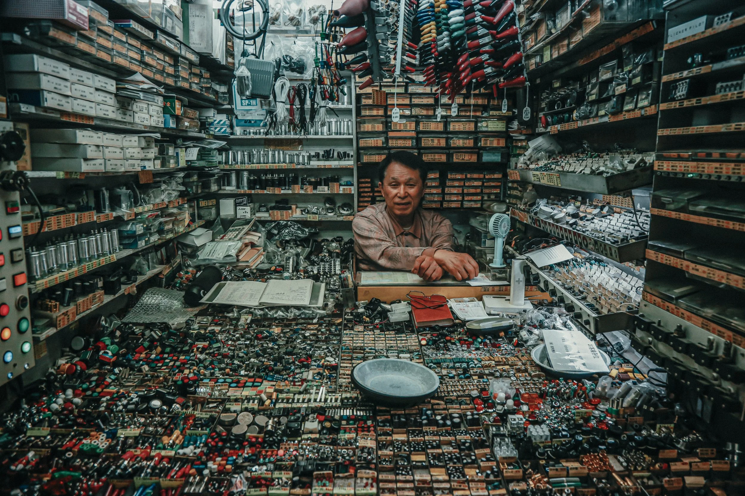 Man zit achter een verkooppui met elektronische onderdelen en schakelaars, omgeven door dozen en losse componenten in een winkel of werkplaats.