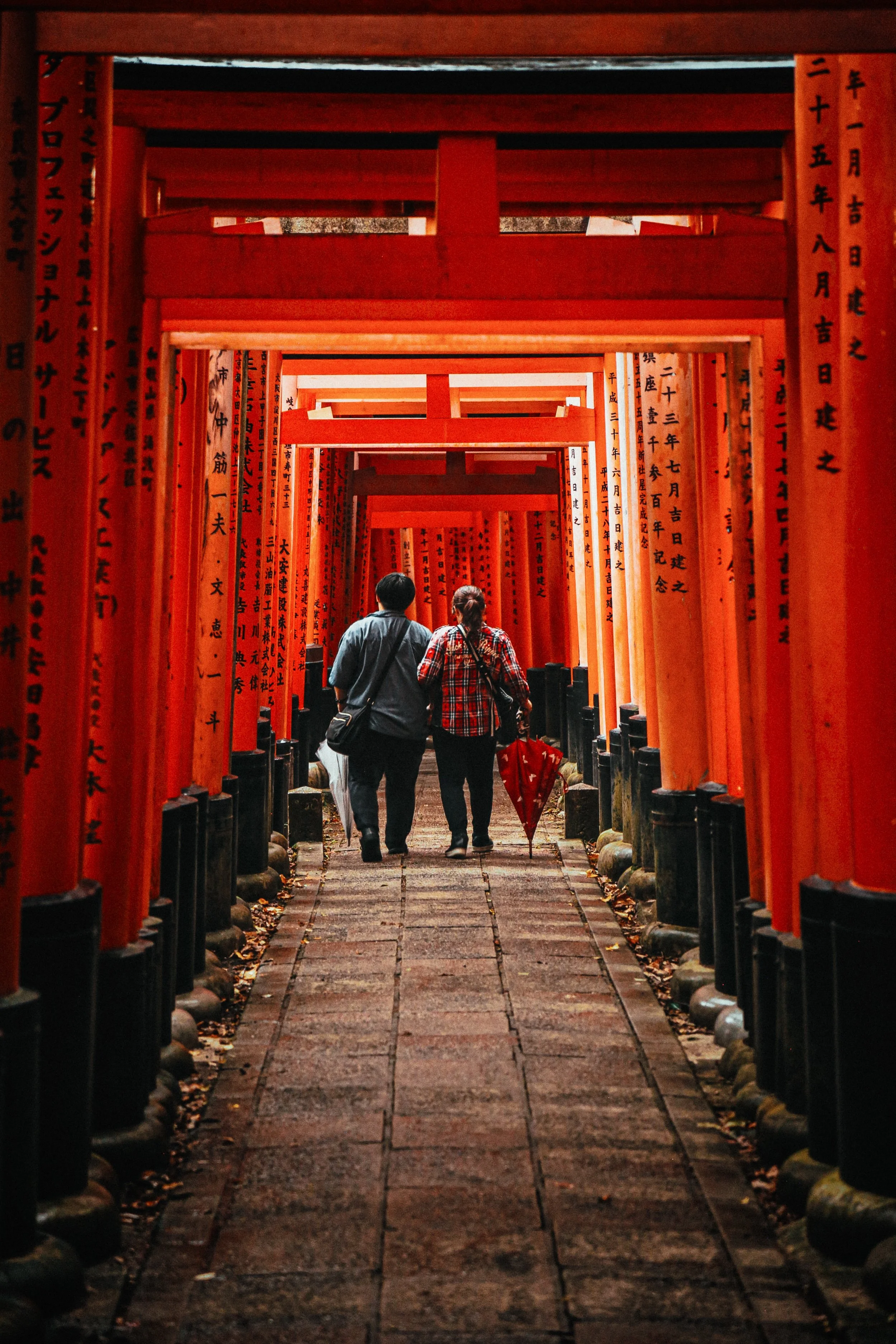 Twee mensen wandelen door een pad omringd door rode torii-poorten met Japanse inscripties, waarschijnlijk in Fushimi Inari-taisha, Kyoto.