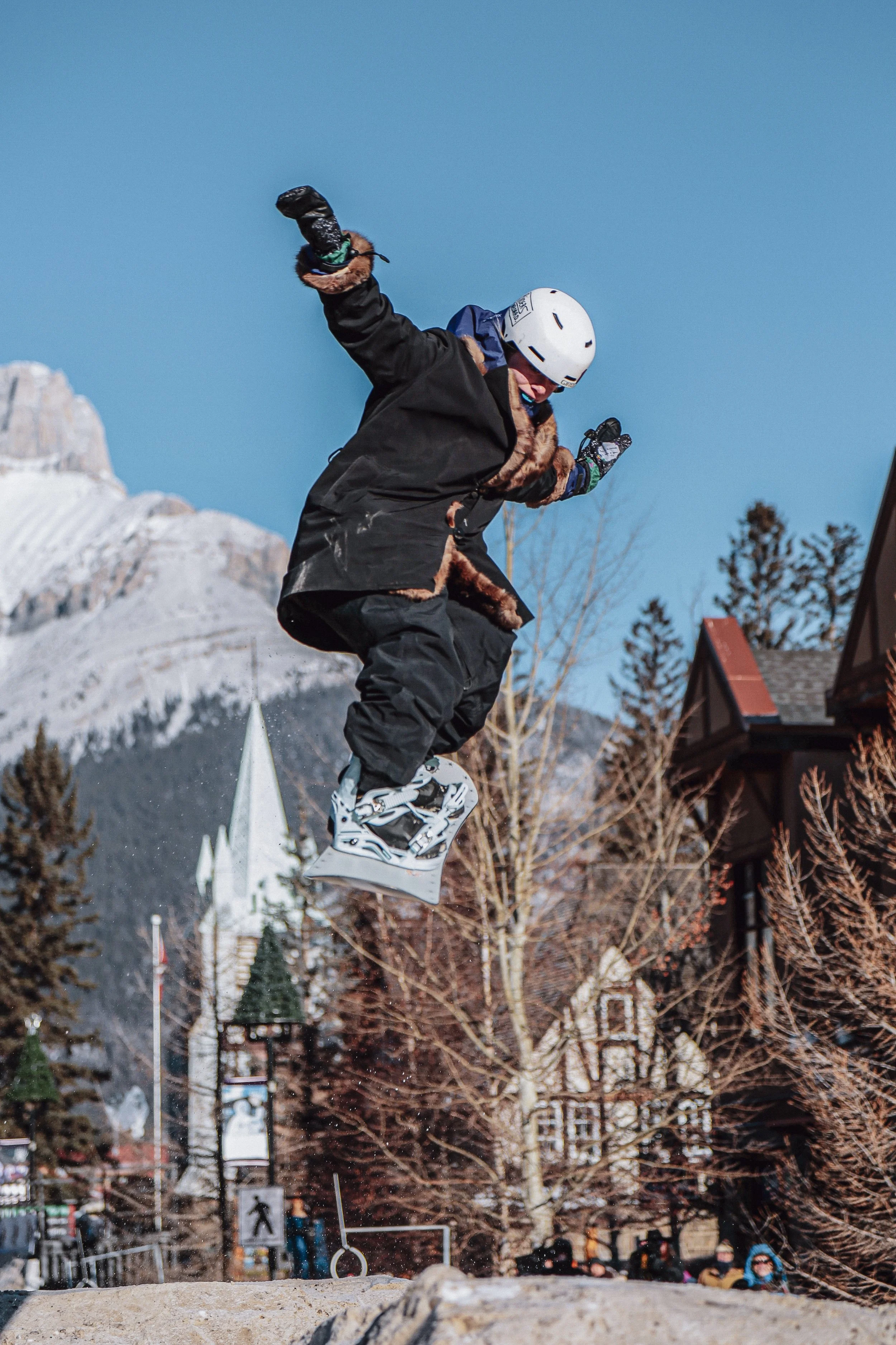 Skiër springt in de lucht op een sneeuwachtige bergachtige achtergrond, draagt een witte helm en een zwarte jas met bontkraag.