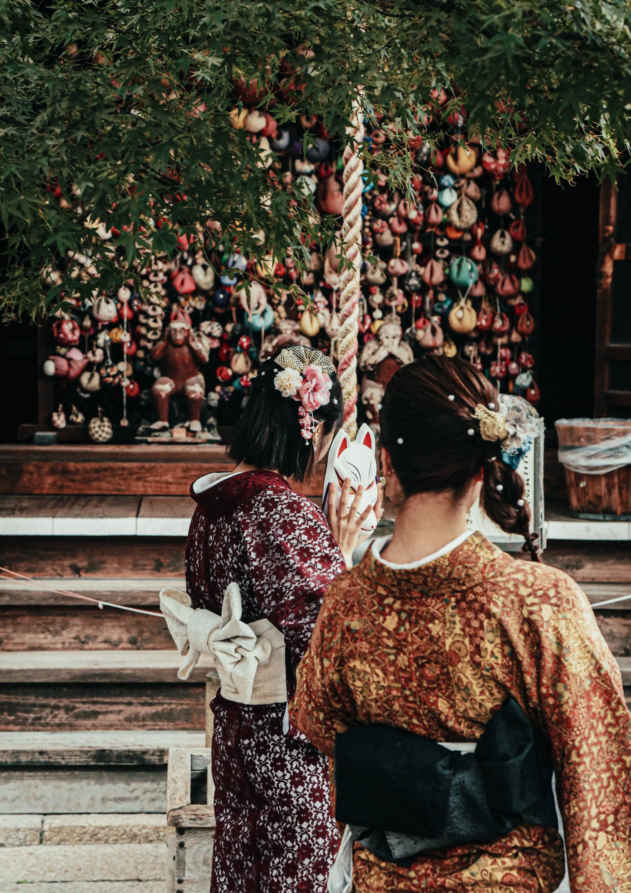 Twee vrouwen gekleed in traditionele Japanse kimono's, één met een katmasker, staan voor een display met kleurrijke kleine voorwerpen en een boom met hangende ornamenten.