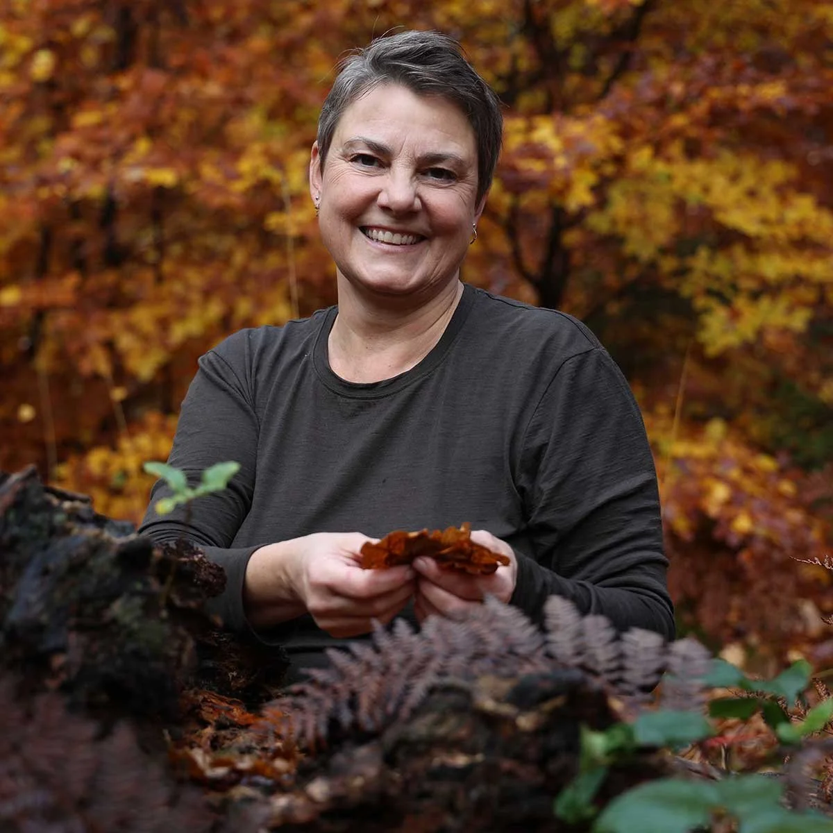 Kvinde med kort hår smiler og holder tørrede blade i en skov med efterårsfarvede blade.