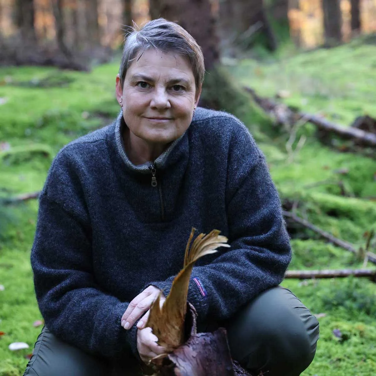 Kvinden i en grøn skov med træer og mosser, holder et stykke bark og inviterer dig til at hjælpe dig selv.