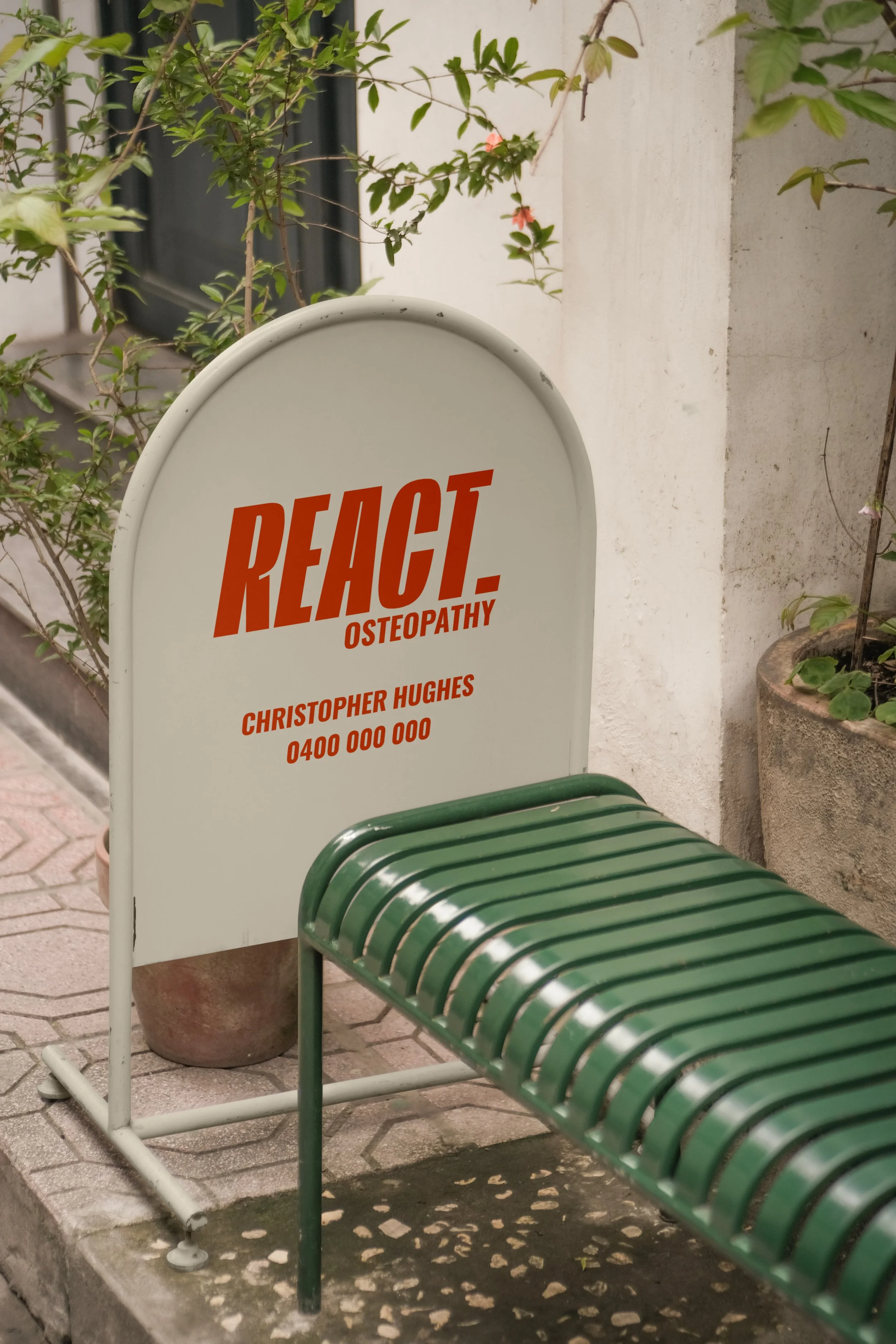 A white signboard with red text advertising REACT Osteopathy, located outdoors on a sidewalk, with potted plants and a green bench nearby.