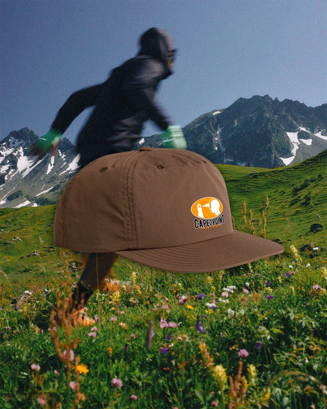 A large, brown cap with a logo reading 'Cape2Point' placed in a field of wildflowers. In the background, a person dressed in black with green gloves is running through a grassy landscape with mountains and snow patches.