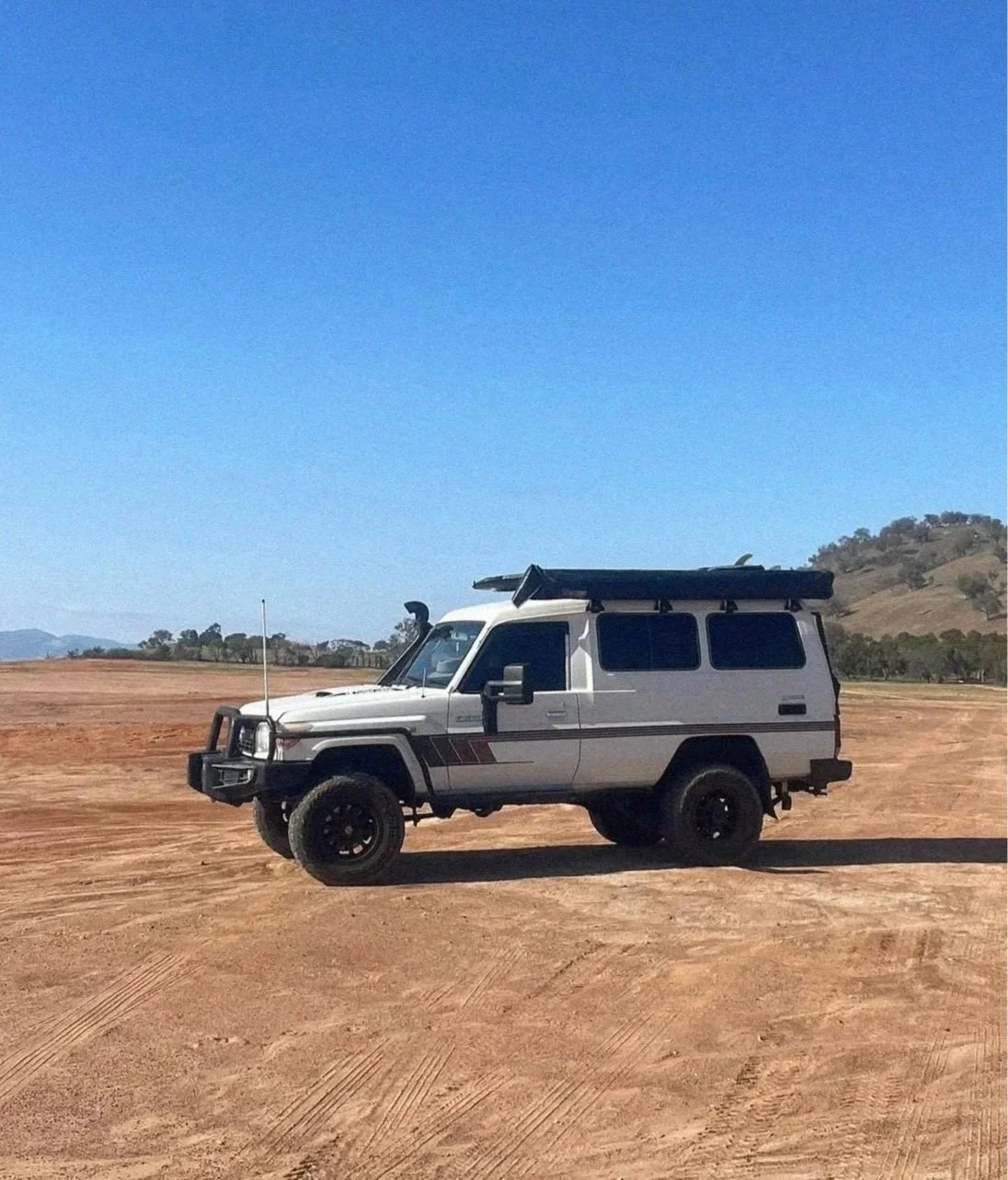 A white off-road vehicle parked on a dirt field under a clear blue sky with distant hills in the background.