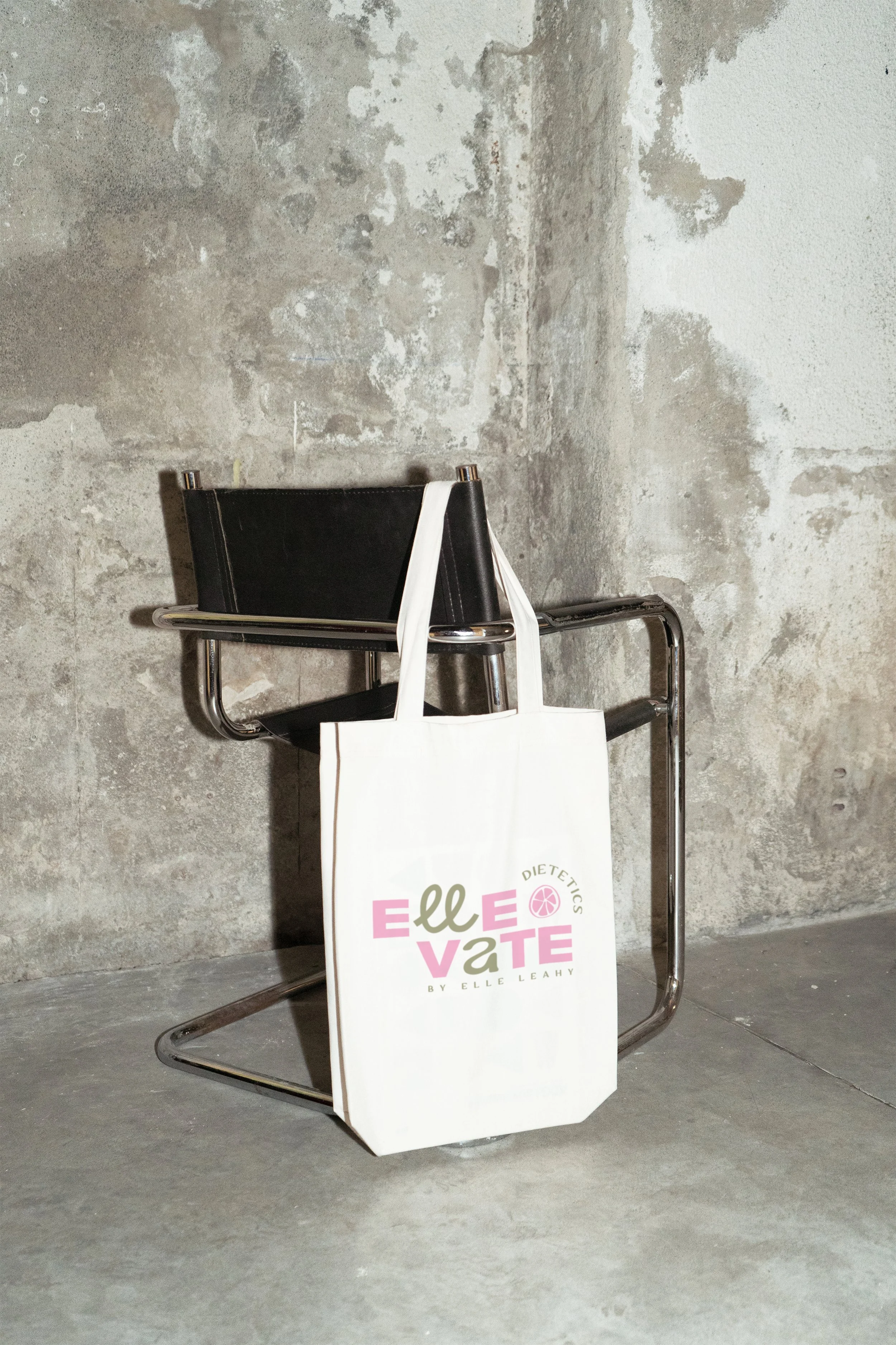 A white tote bag hanging on a chrome chair with a black seat, placed against a concrete wall.