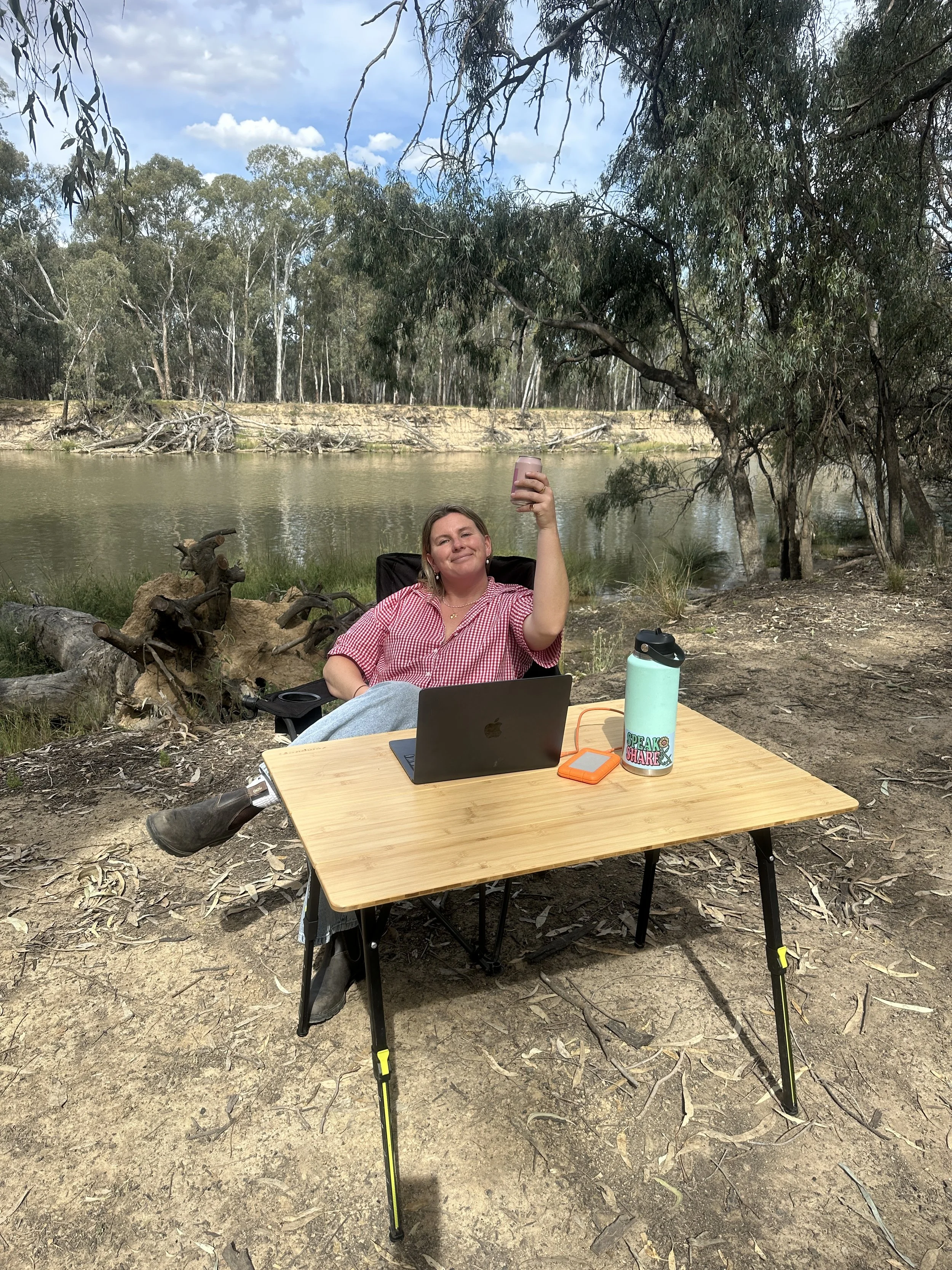 A woman sitting at an outdoor table near a river, holding a smartphone up in a toast gesture and smiling. The table has a laptop, a water bottle, and a small electronic device. She is surrounded by trees and natural landscape.