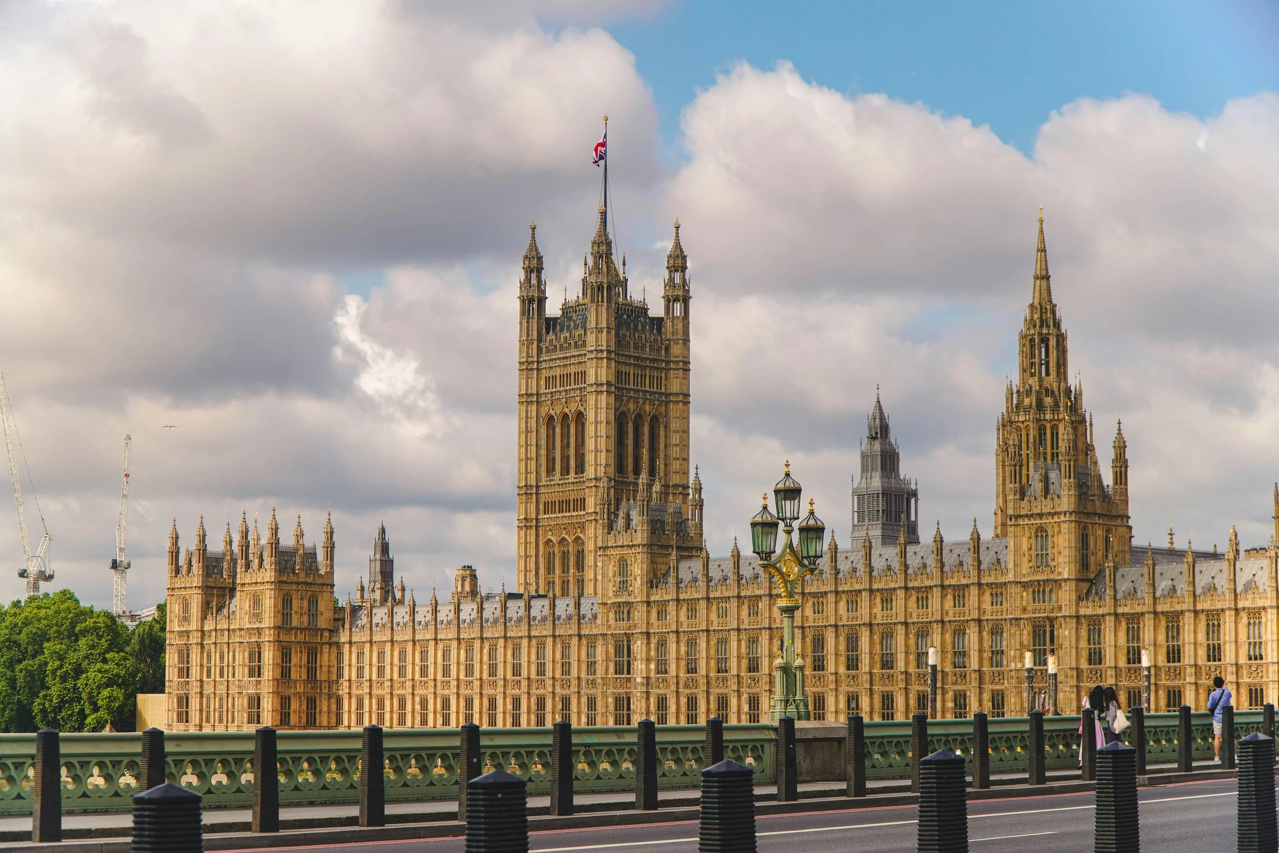 View of the Palace of Westminster in London, showing its gothic architecture and clock tower, with traffic lights and pedestrians in the foreground under a partly cloudy sky.