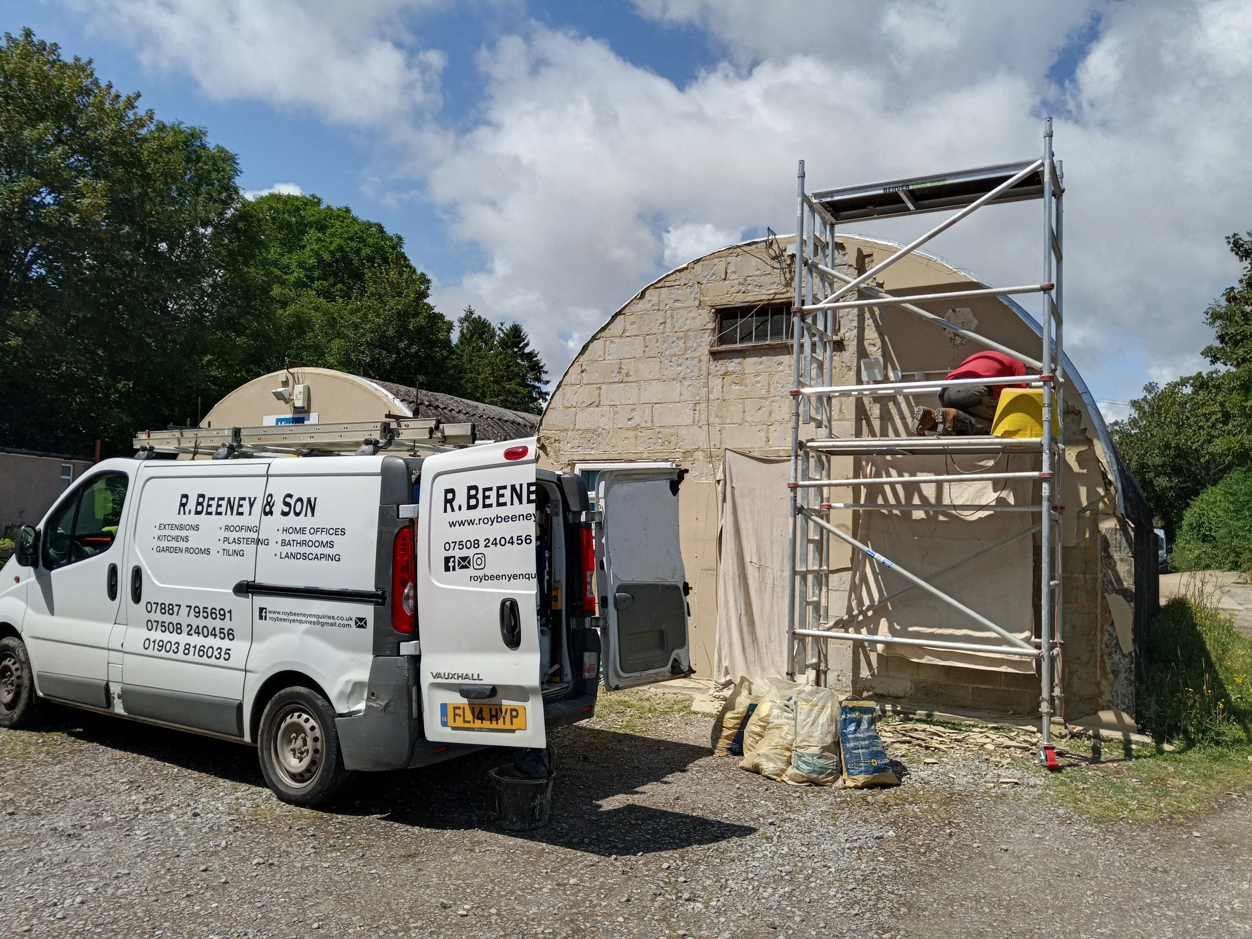 Construction workers repairing the exterior of a small building with scaffolding, next to a company van parked on a gravel surface.