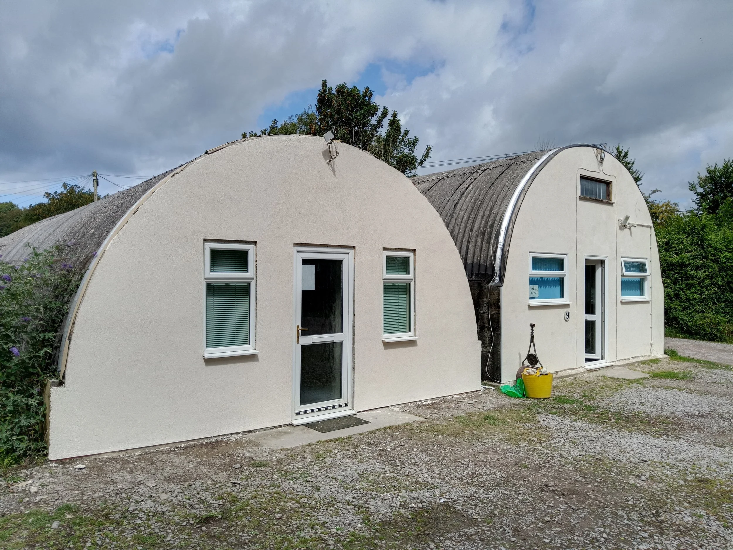 Two Quonset hut houses with white exteriors and arched roofs, situated outdoors with some greenery in the background, cloudy sky overhead, and gardening tools and a yellow bucket in front.