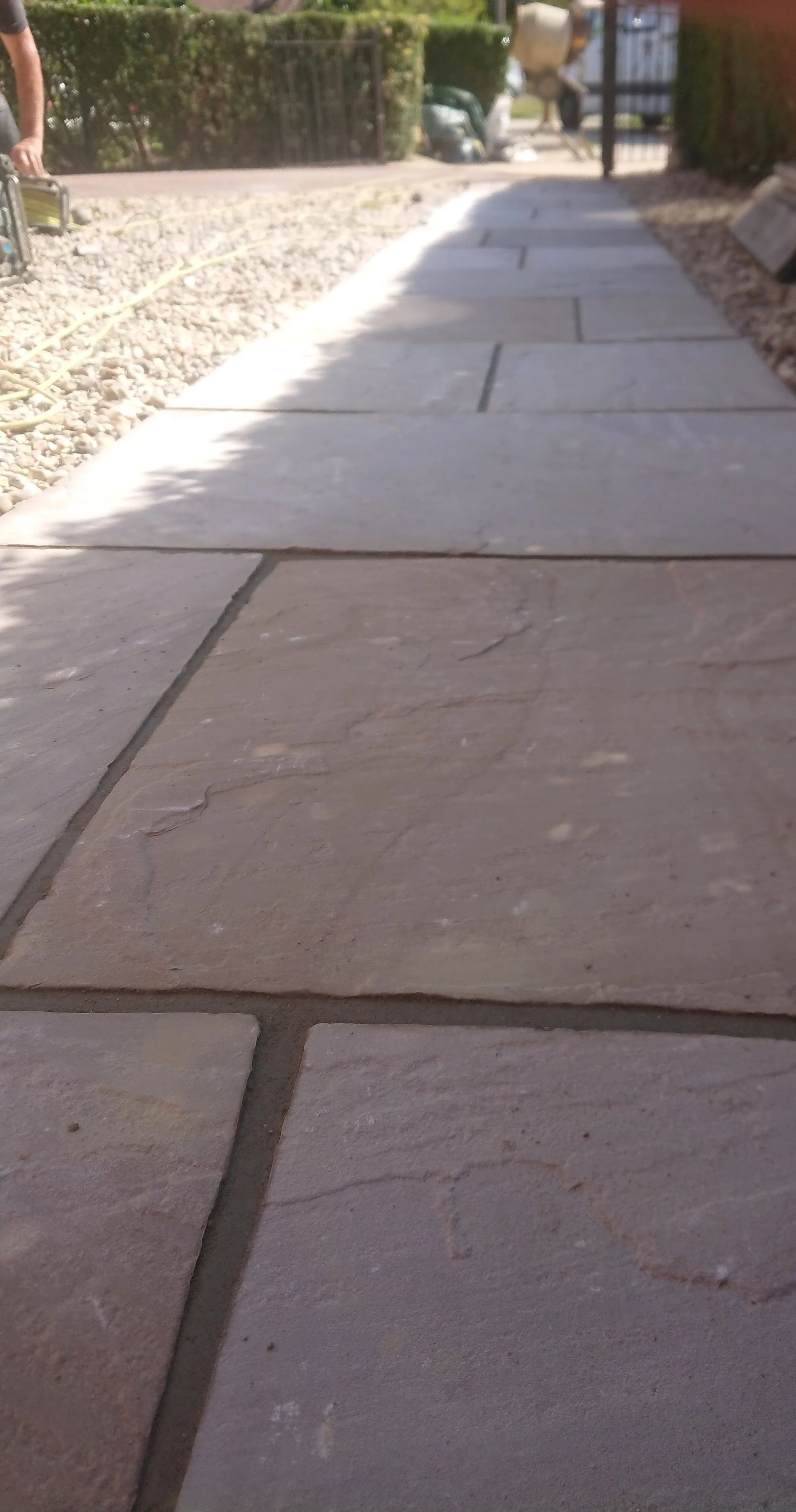 Close-up view of a stone walkway with rectangular pavers, with some gravel and plants along the side, and a cinder block wall in the background.