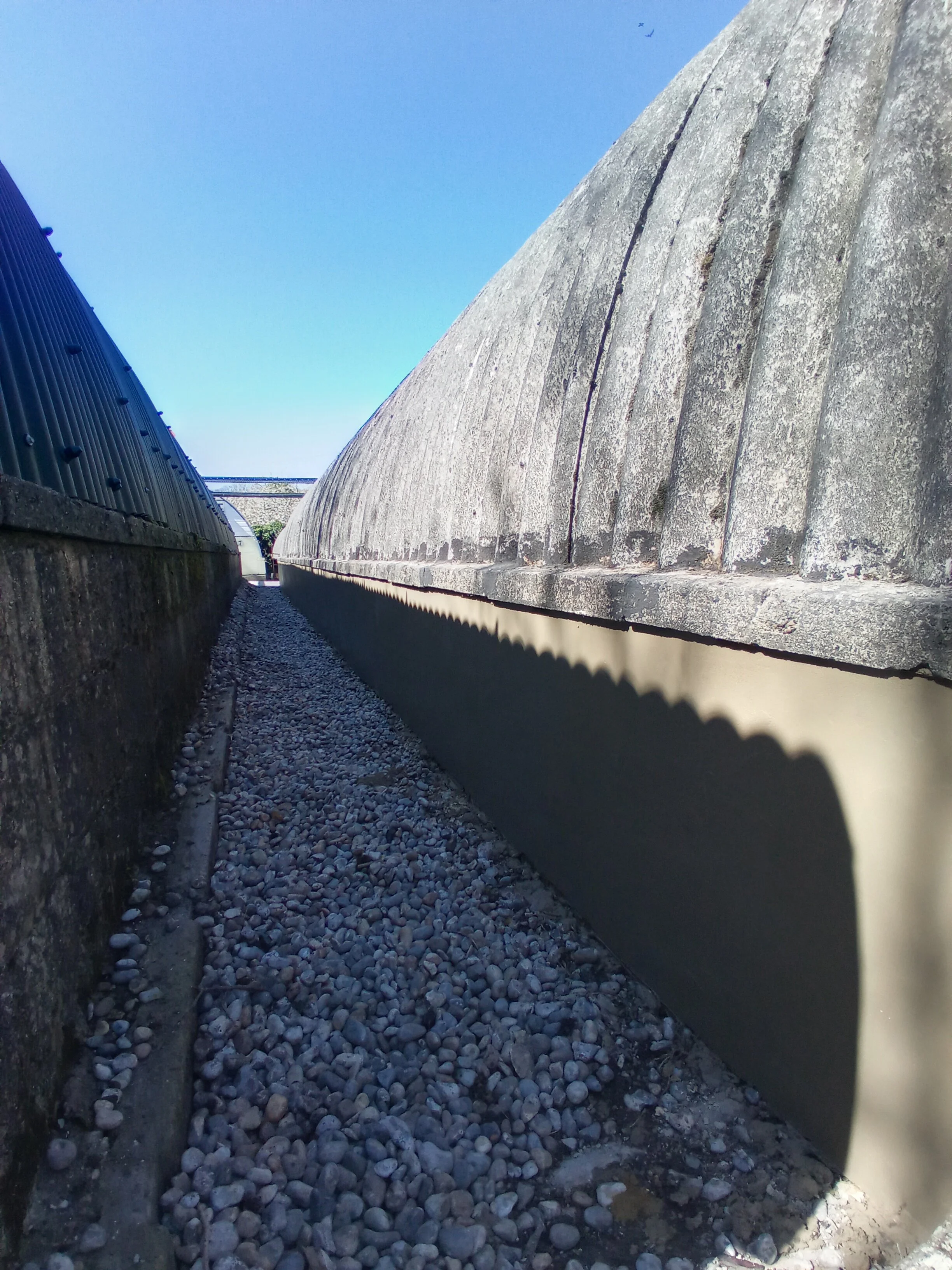 View of two large concrete tanks or silos, one with a blue metal exterior and the other with a weathered gray surface, separated by a narrow gravel pathway, under a clear blue sky.