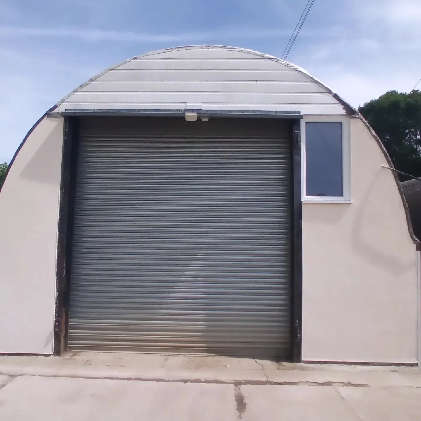 A small building with a curved roof and a large metal roll-up door, with a small window to the right of the door, set on a concrete ground under a blue sky.