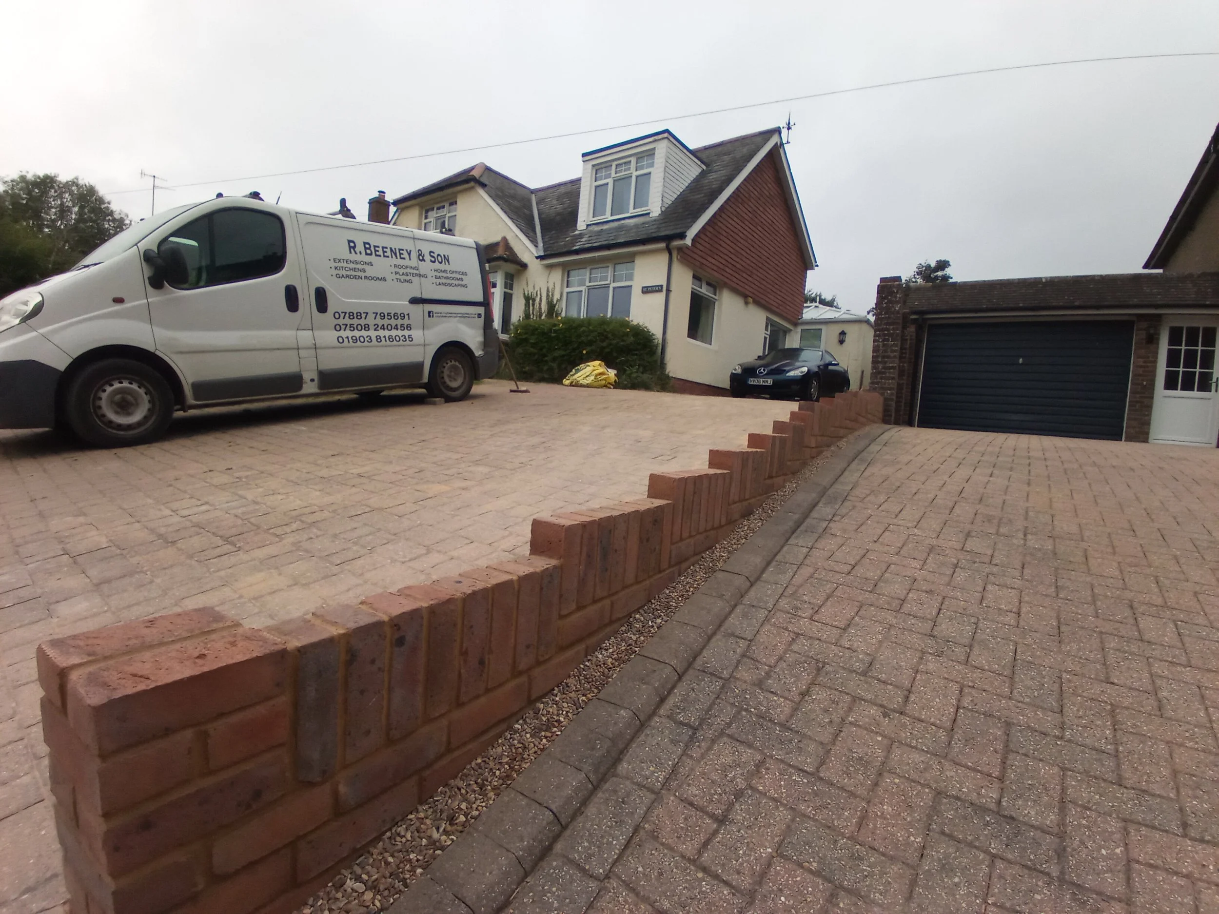 Residential driveway with paved brick and concrete surface, a brick wall edge, a white construction van parked, and a house with a garage in the background.