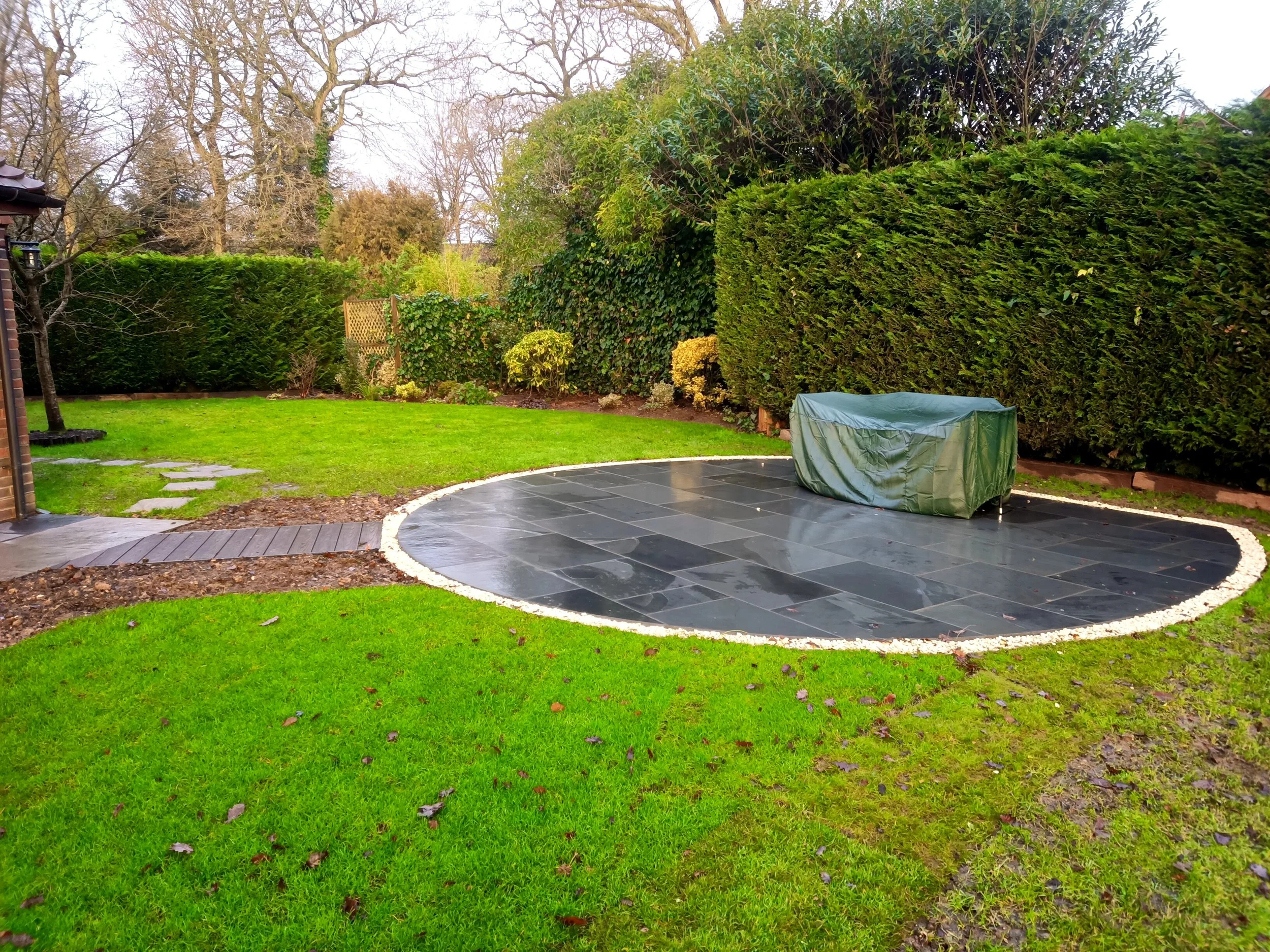  Garden with green grass, Slate slab circular patio area with a covered object, and a tall hedge along the perimeter.