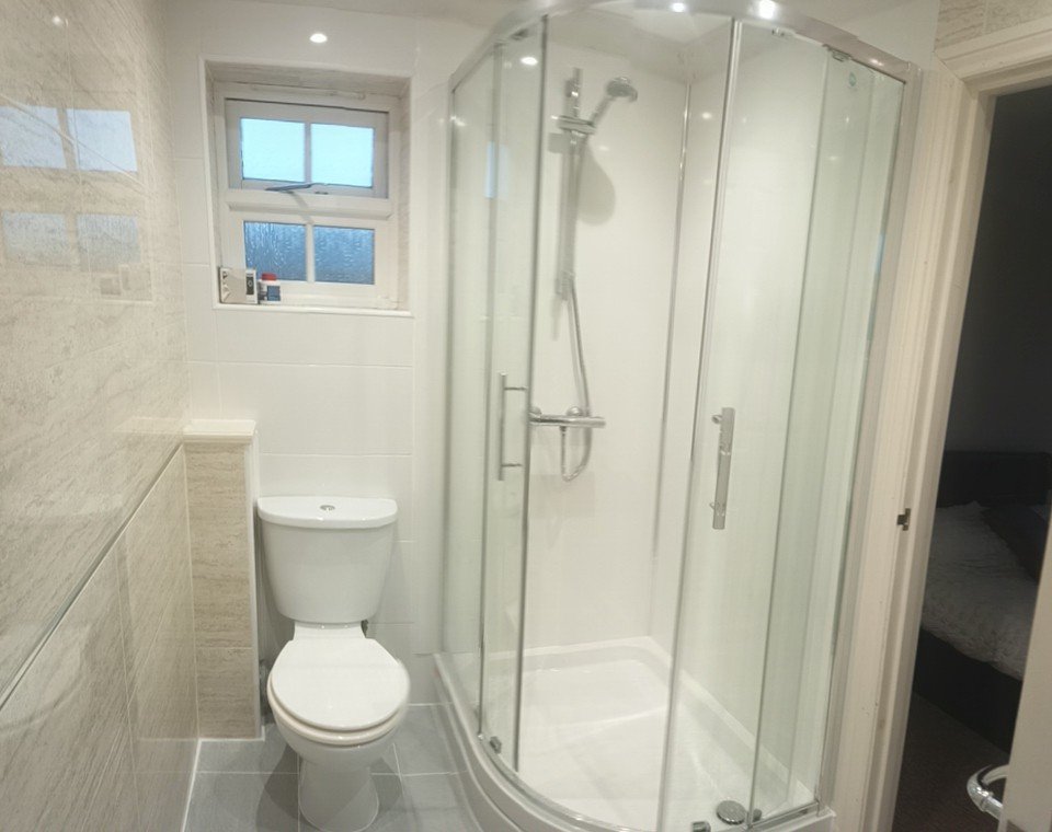 Small bathroom with a toilet, a shower with glass doors, a small frosted window, and a glimpse into a bedroom.