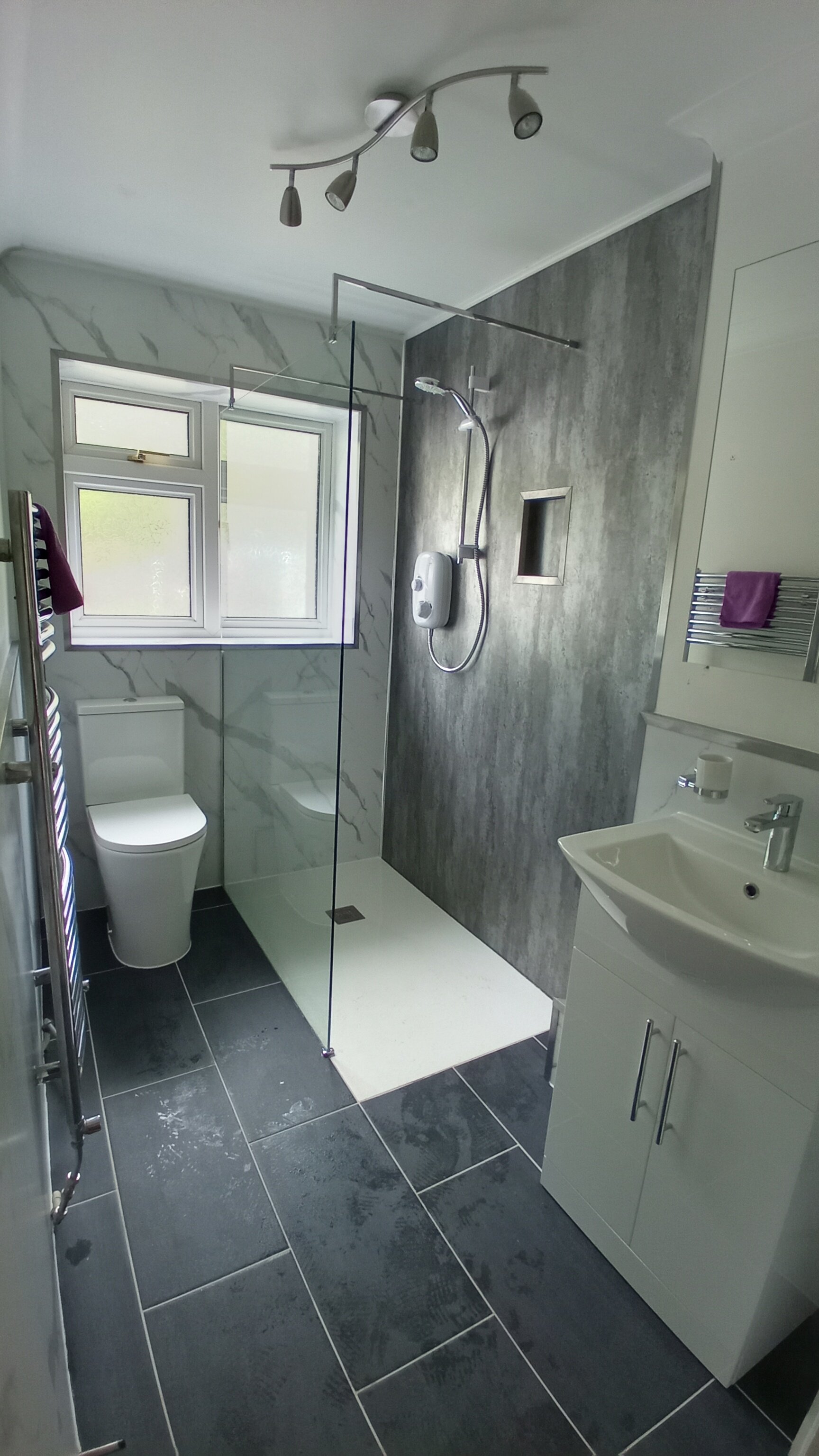 Modern bathroom with walk-in shower featuring gray wall and floor tiles, a white toilet, a white vanity with sink, a window, and a towel rack.