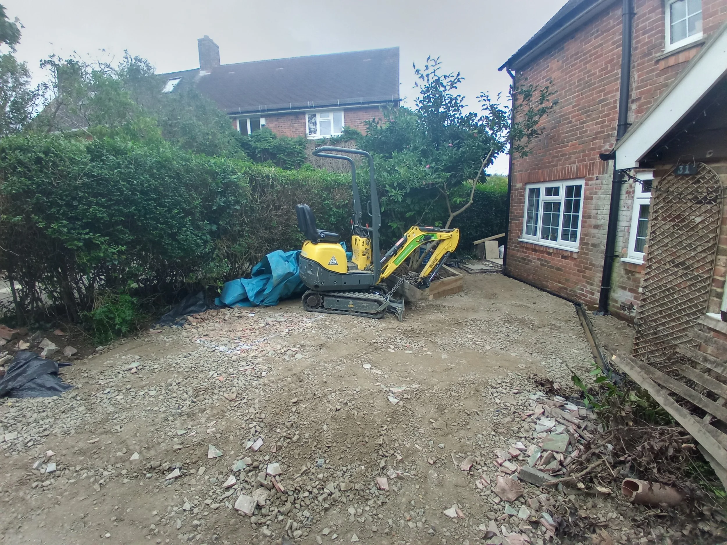 A construction site in a backyard with a small yellow excavator, dirt, and debris, surrounded by a brick house, a garden hedge, and some foliage.