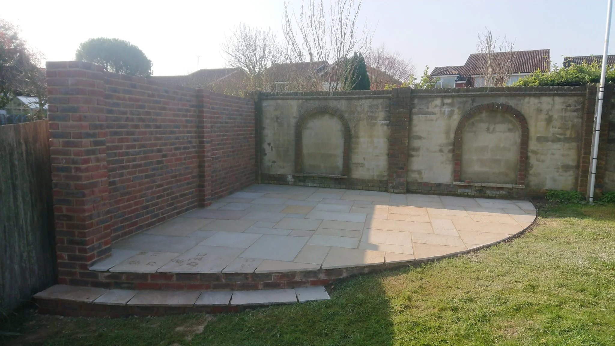 A backyard patio with a newly laid stone surface, bordered by a brick and concrete wall, with some grass and a curved edge.