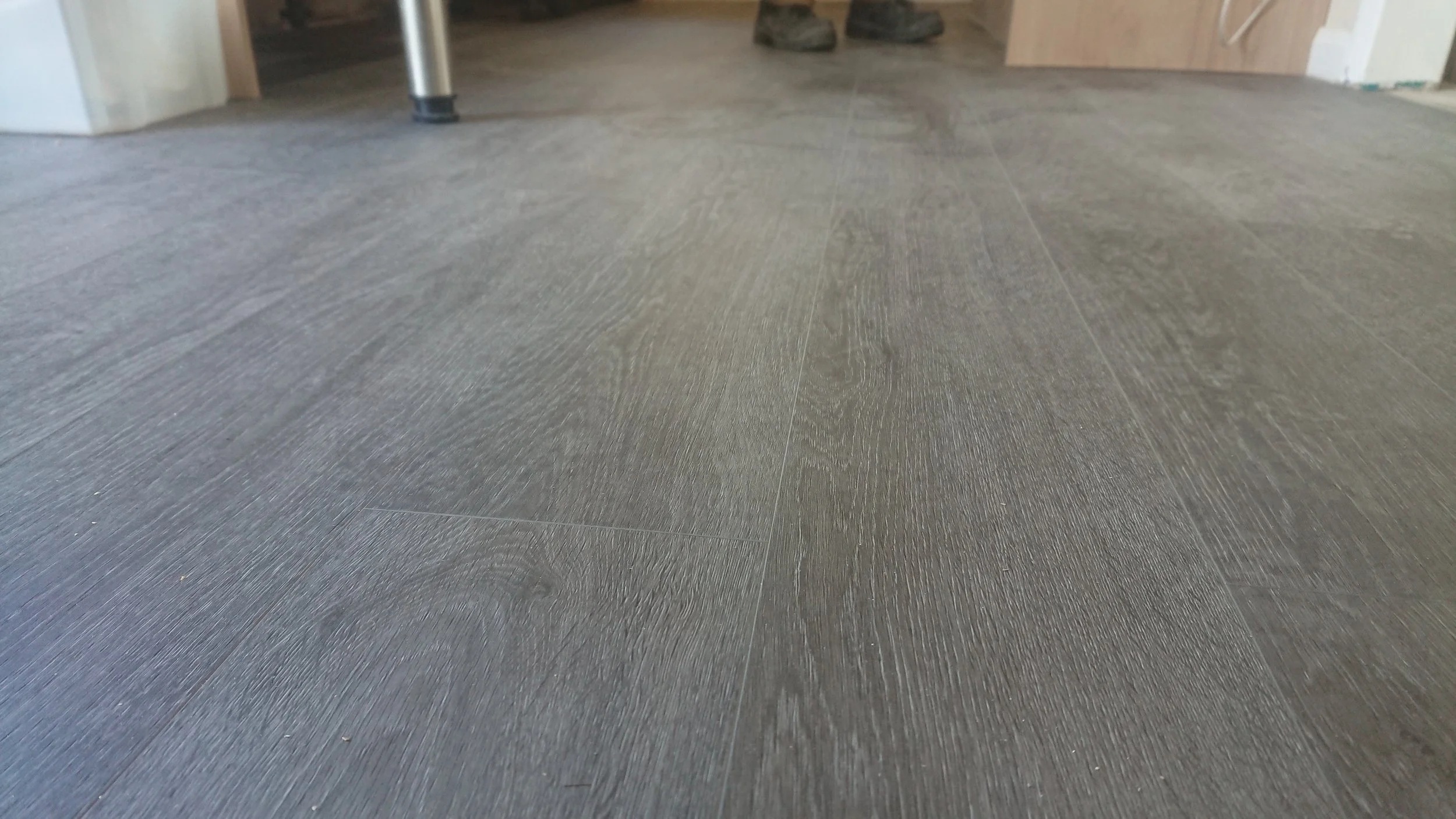 Close-up of a dark wood floor with visible grain patterns, with part of a metal leg of a piece of furniture and some shoes visible in the background.