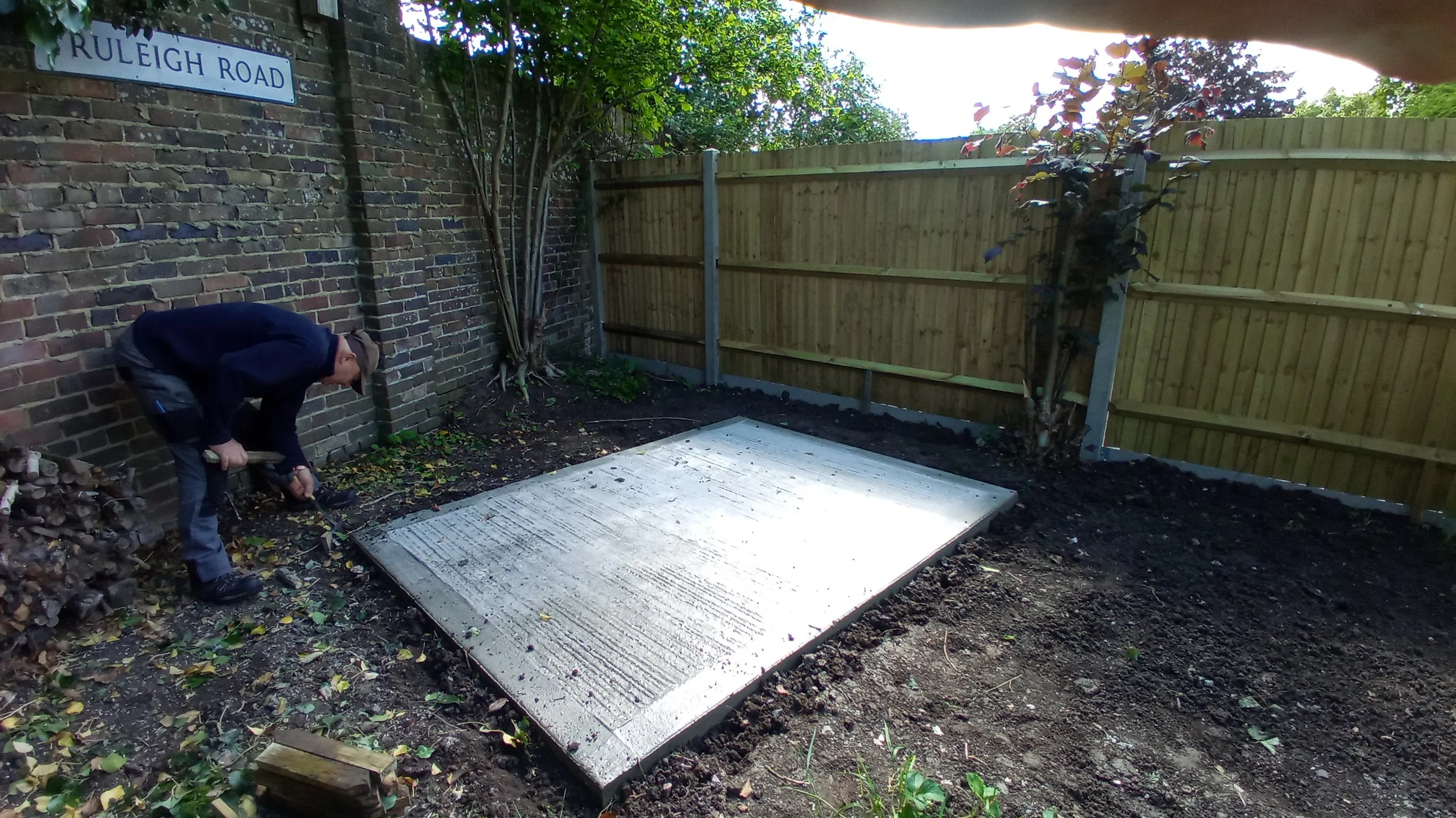 A man working on a concrete slab in a back garden, with a brick wall on the left, a fence on the right, and some trees and plants in the background.