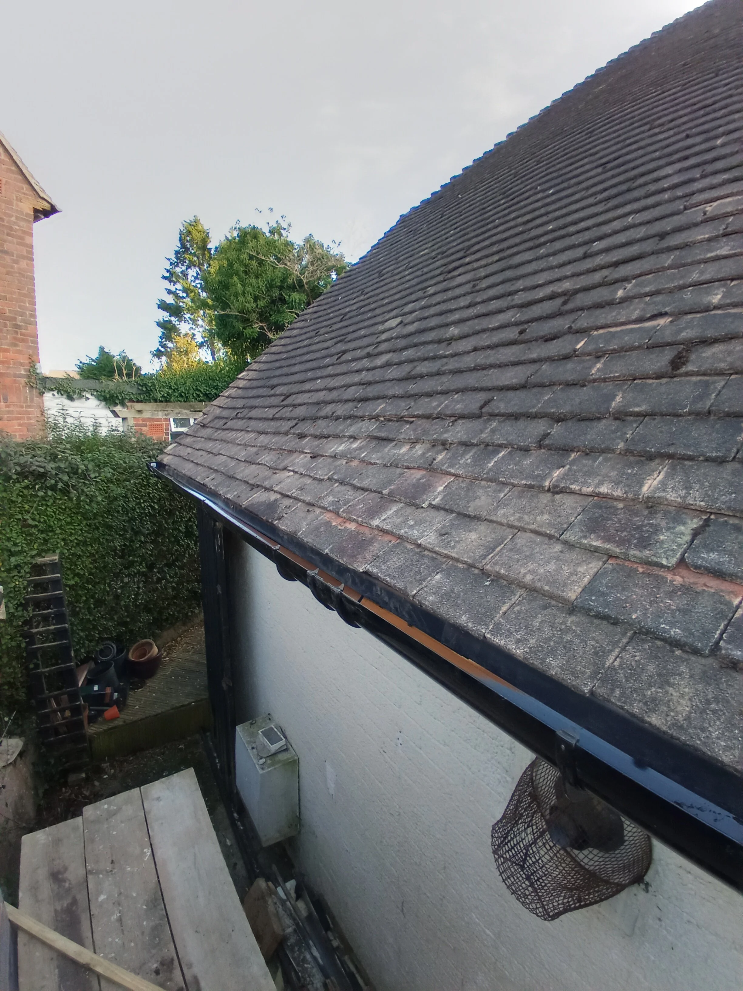 View of a residential house roof with brown tiles, a white exterior wall, and a small outdoor area with a wooden platform, plants, and gardening tools.